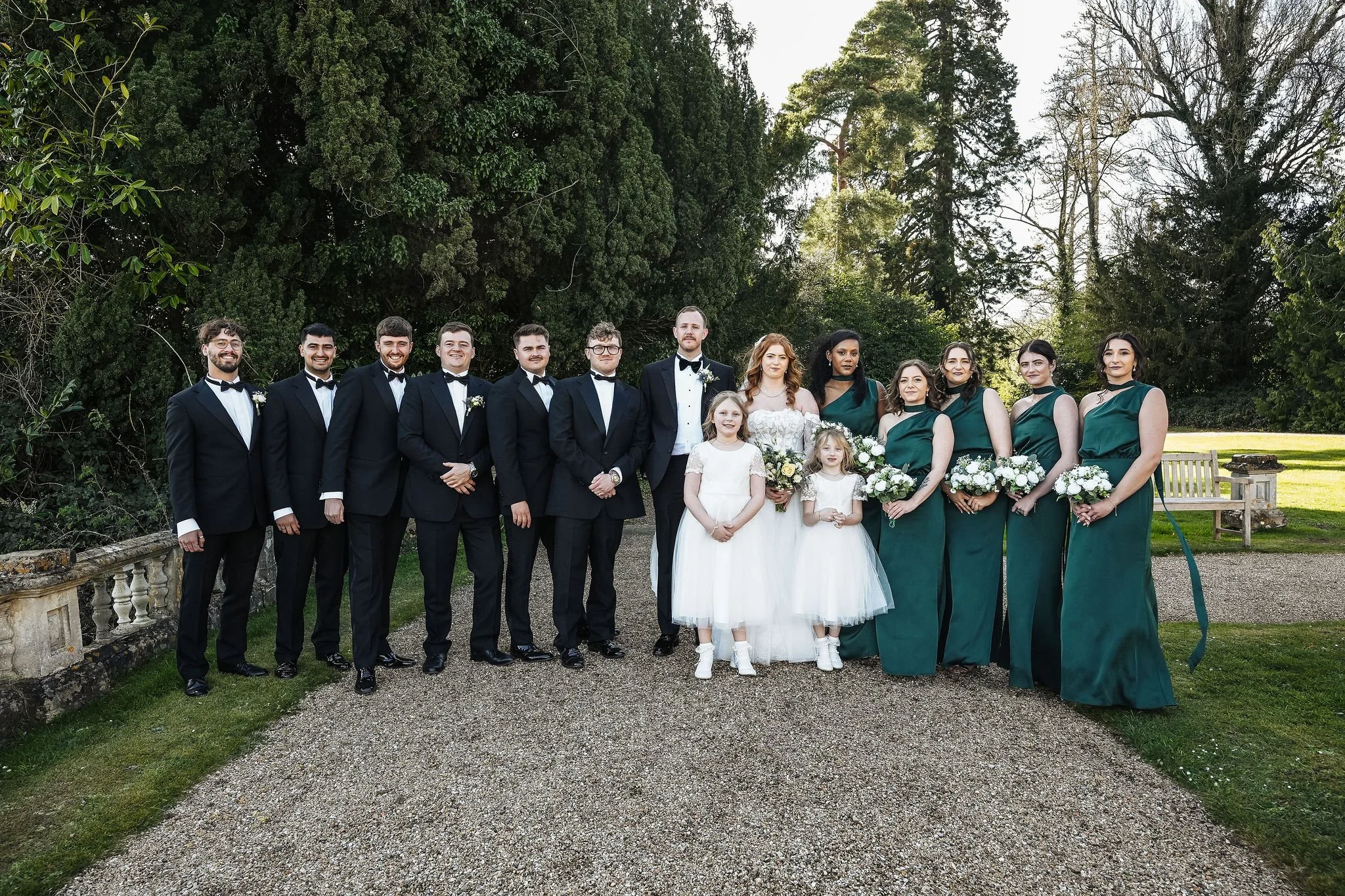 Group of people dressed in formal attire, including wedding dresses, tuxedos, and bridesmaid dresses, standing outdoors in front of large trees and greenery.