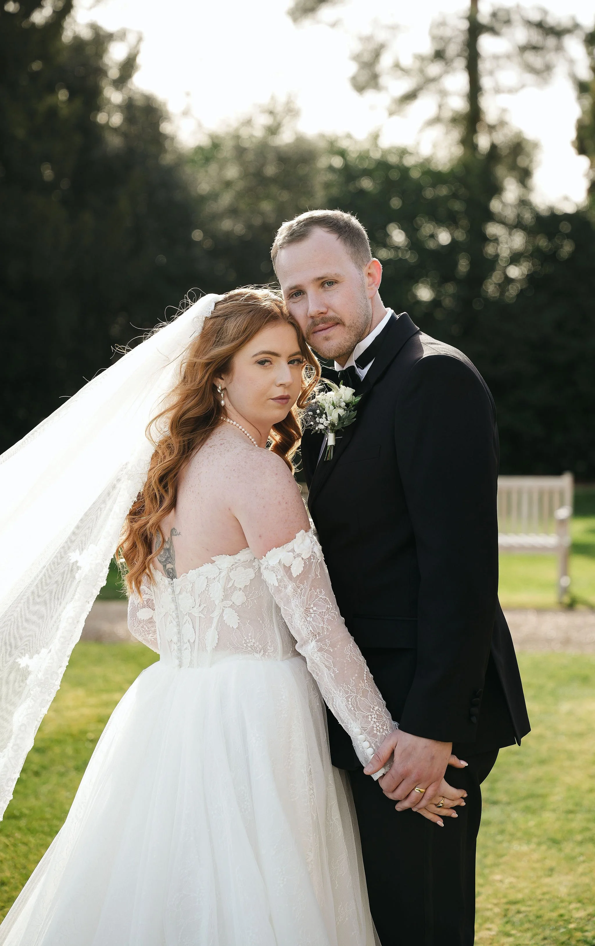 A bride with long, wavy red hair and a groom with short light hair standing outdoors, holding hands, with trees and a bench in the background at sunset.