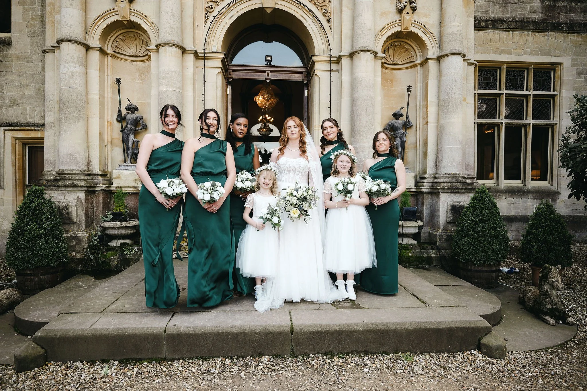 A group of seven women and two young girls standing on a stone step in front of a historic building with large columns and statues, participating in a wedding celebration. The women are dressed in green dresses, and the bridesmaids are holding bouque