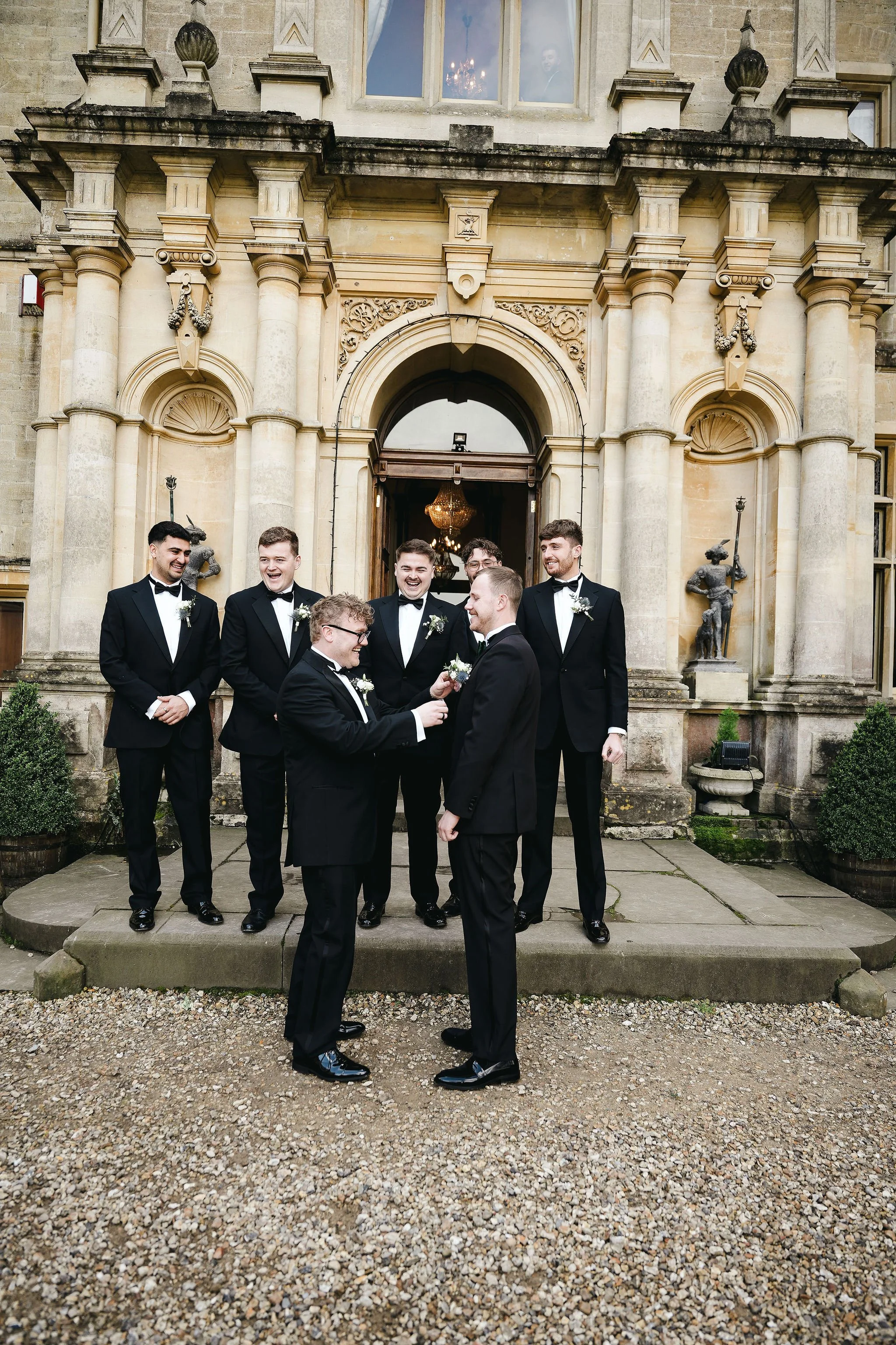 A group of men dressed in tuxedos standing outside a historic stone building with ornate architecture, columns, and statues, possibly for a wedding.