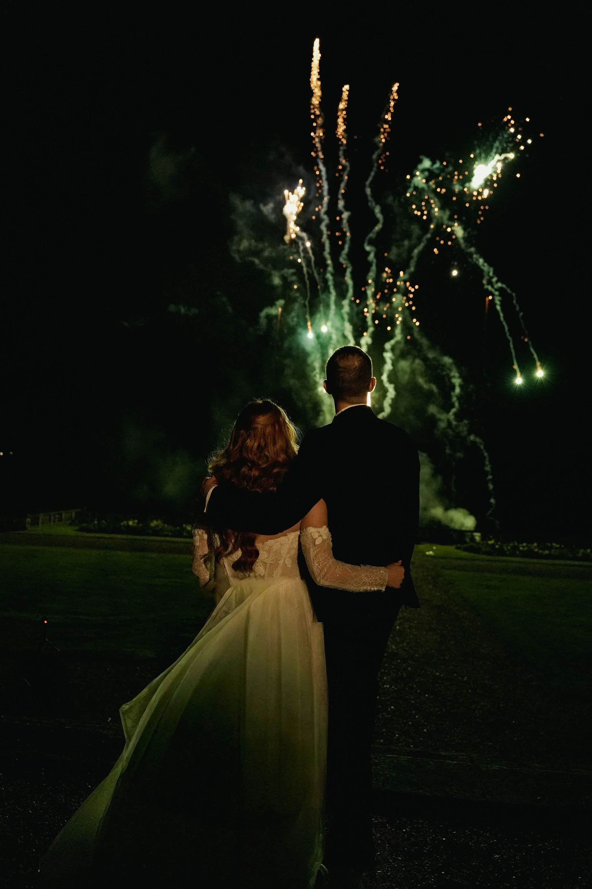 A newlywed couple, a woman in a wedding dress with long curly hair and a man in a dark suit, embraces while watching fireworks in the night sky. They are standing outdoors on a grassy area.