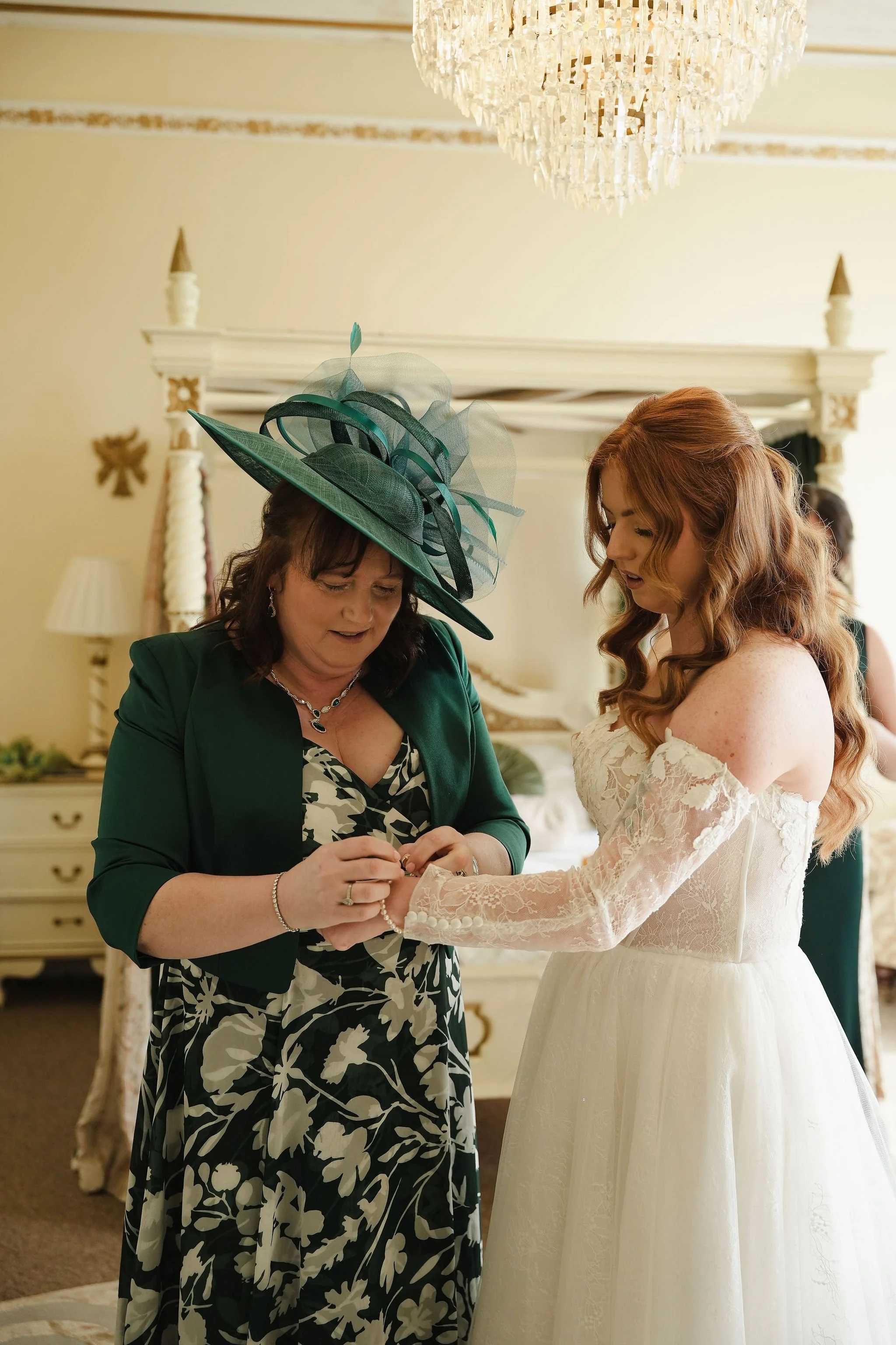 A woman with a large green and blue hat placing a ring on a bride's finger during a wedding ceremony in an elegant room with a chandelier