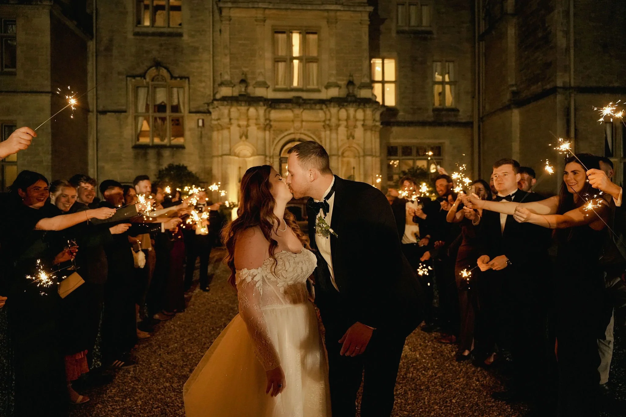 A newlywed couple kissing at night during a wedding celebration, surrounded by guests holding sparklers, with a historic building in the background.