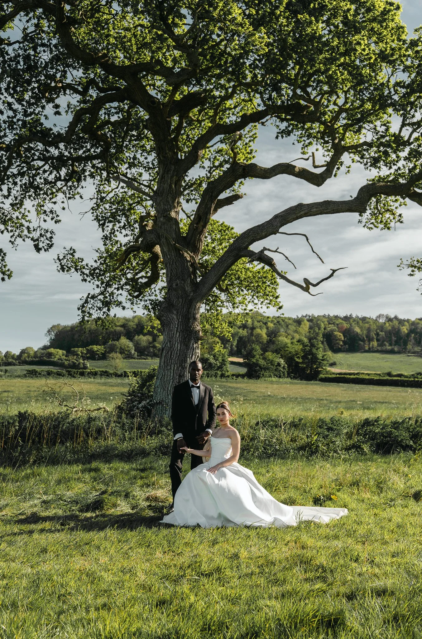 A man in a black tuxedo and a woman in a white wedding dress sitting in a grassy field under a large, leafless tree with sprawling branches. The background features rolling hills and a partly cloudy sky.