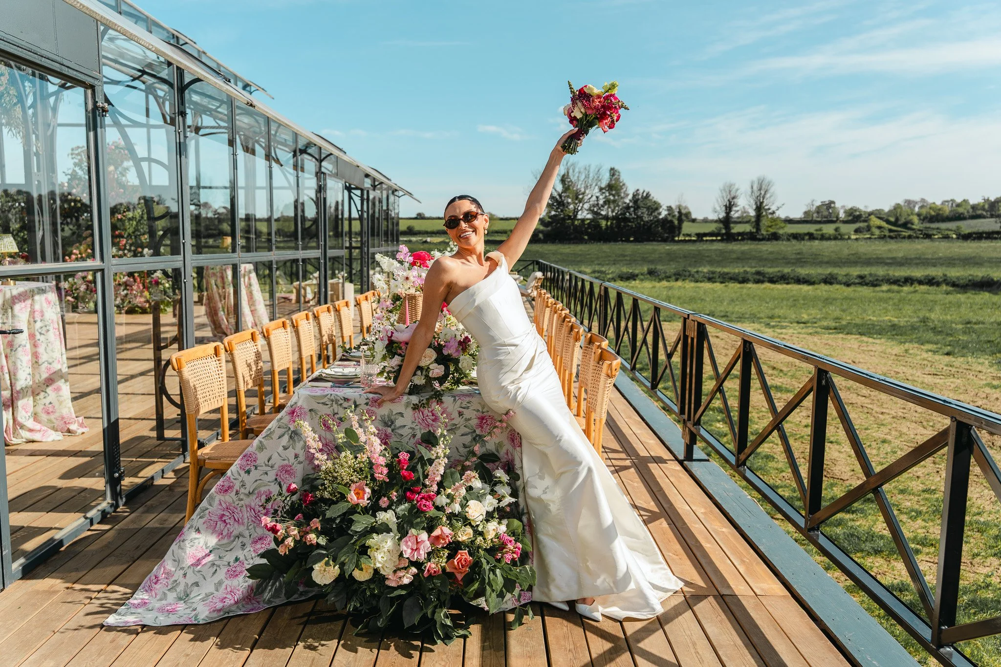 A woman in a white dress and sunglasses holding a bouquet of flowers high above her head, smiling, standing on an outdoor wooden terrace decorated with a floral tablecloth and a long flower arrangement, with a background of green fields and a clear b