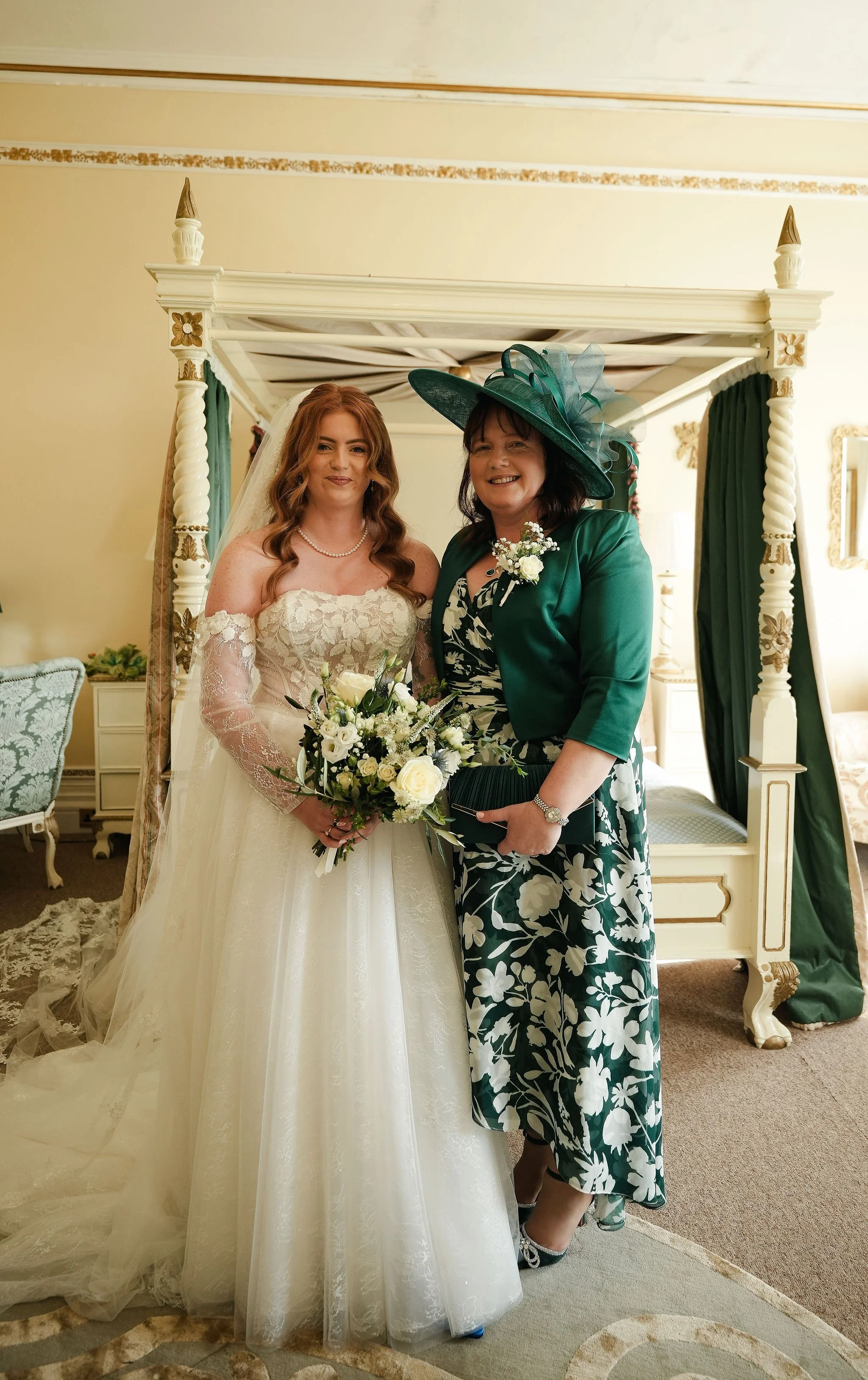A bride in a white wedding gown holding a bouquet stands next to a woman in a green floral dress and matching hat inside a decorated room with a canopy bed.