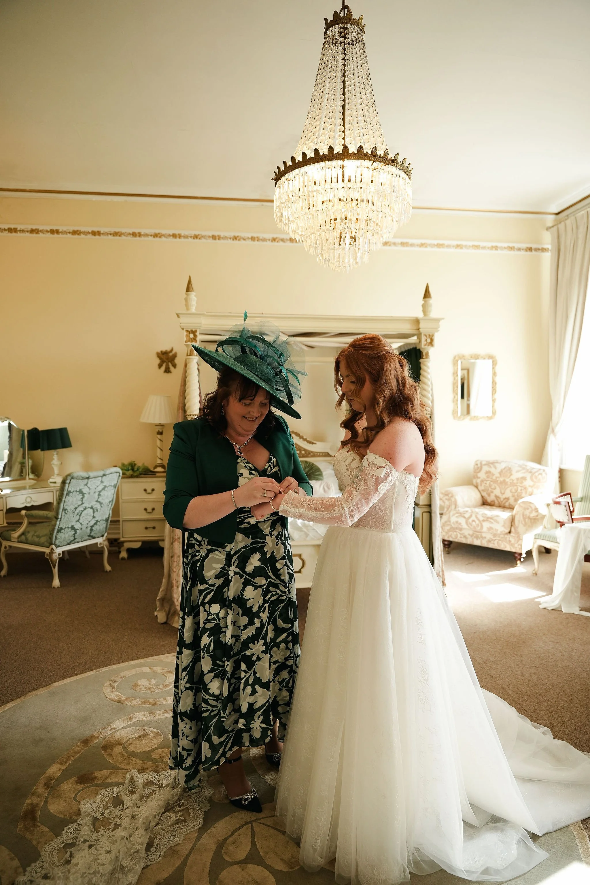 A bride and an older woman are standing in a well-lit, elegant room with a chandelier hanging from the ceiling. The older woman is helping the bride with her wedding dress. The room has vintage furniture, a large mirror, and cream-colored walls.