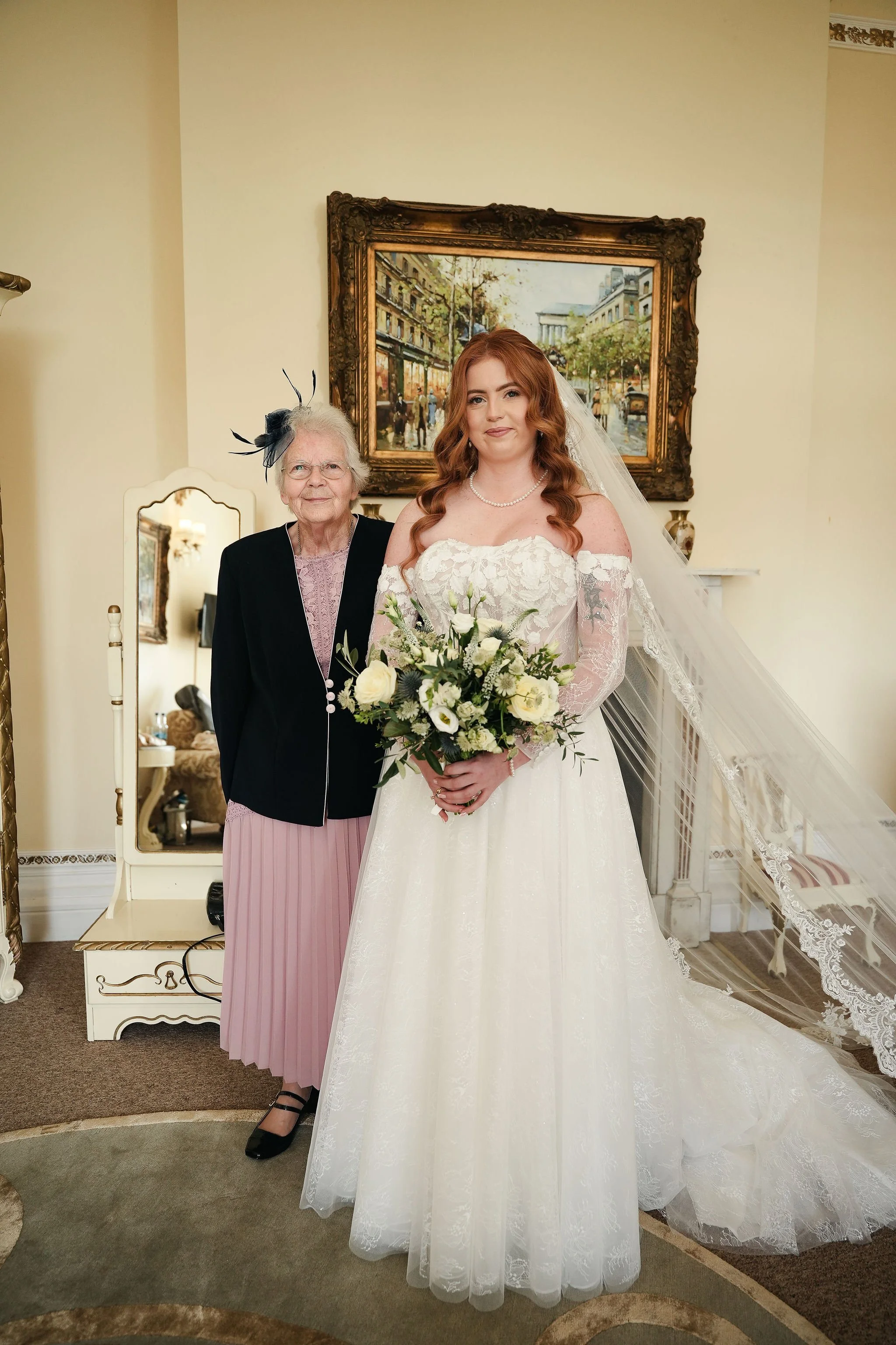 A bride in a white wedding gown holding a bouquet of white flowers, standing next to an elderly woman dressed in a black jacket, pink dress, and a fascinator, in a room with a painting of a city street and a mirror.