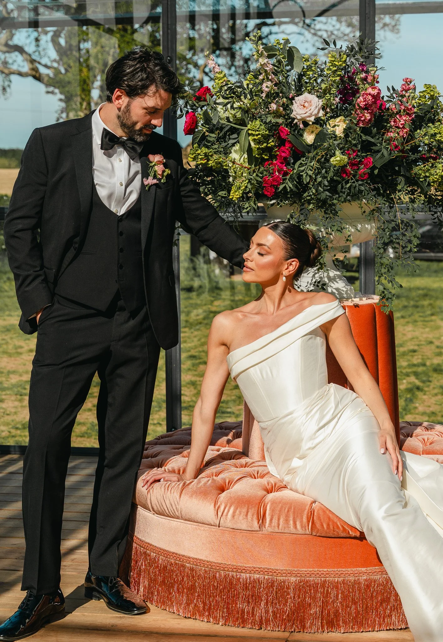 A man in a tuxedo with a boutonniere stands beside a woman in a white dress, who is seated on an elegant, vintage pink velvet couch with fringe, outdoors in front of a floral arrangement, during daytime.