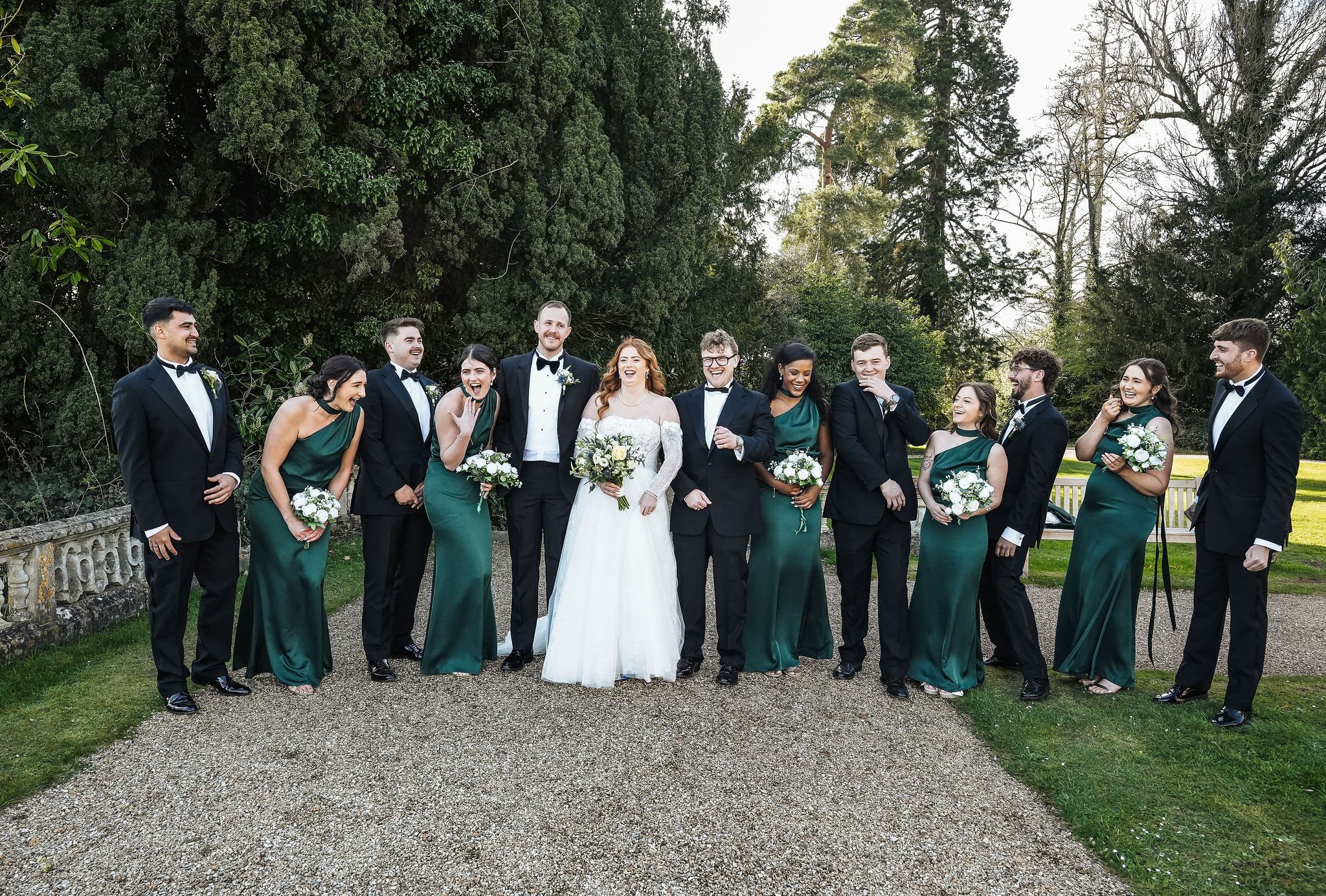 Group of people dressed in formal attire, with some wearing tuxedos and others in green dresses, standing outdoors in front of a background of tall trees and greenery, smiling and laughing, holding bouquets, celebrating a wedding.