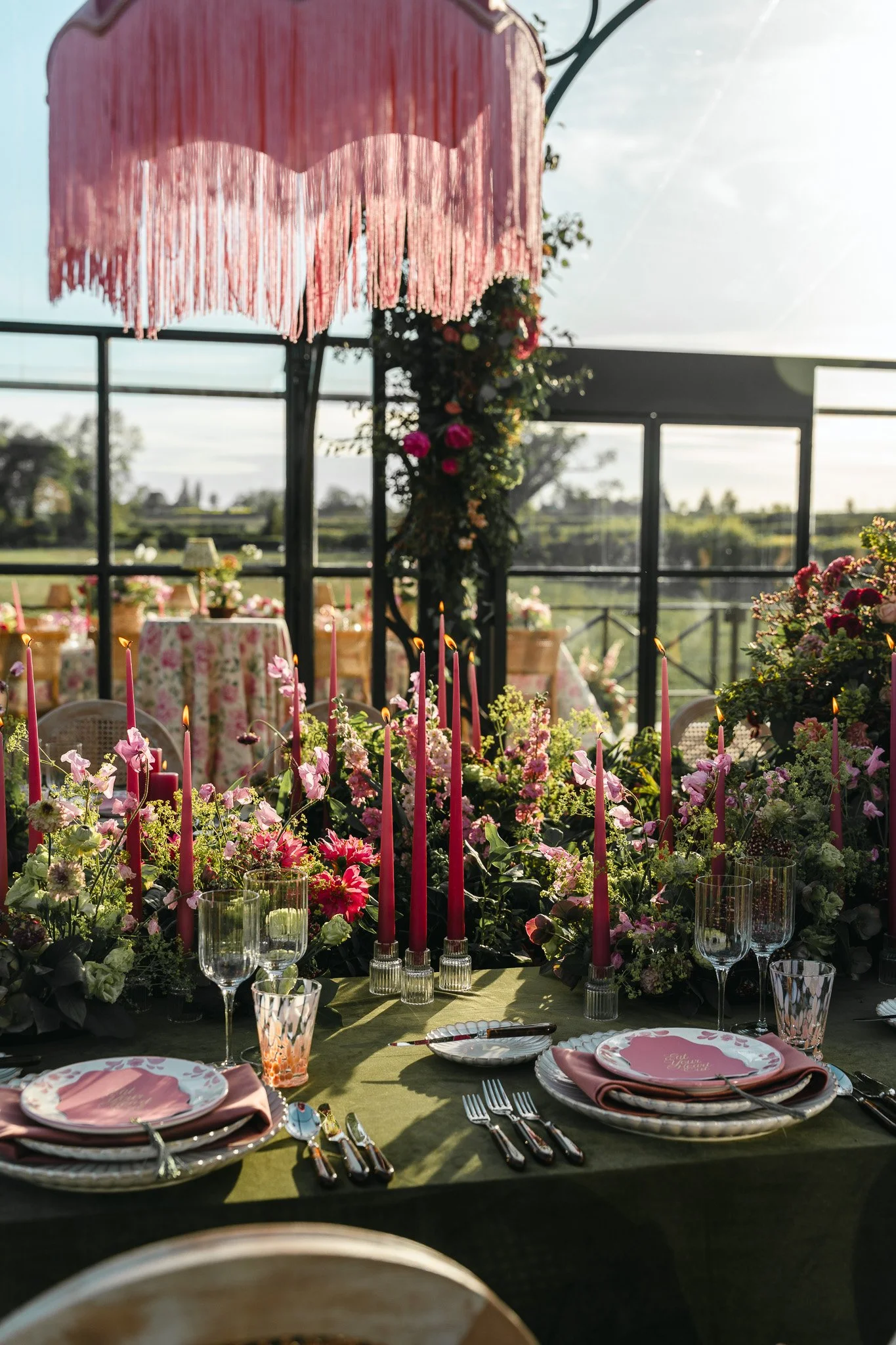 Elegant wedding reception table decorated with pink candles, pink and white flowers, and matching pink plates, with a floral backdrop and outdoor scenery.