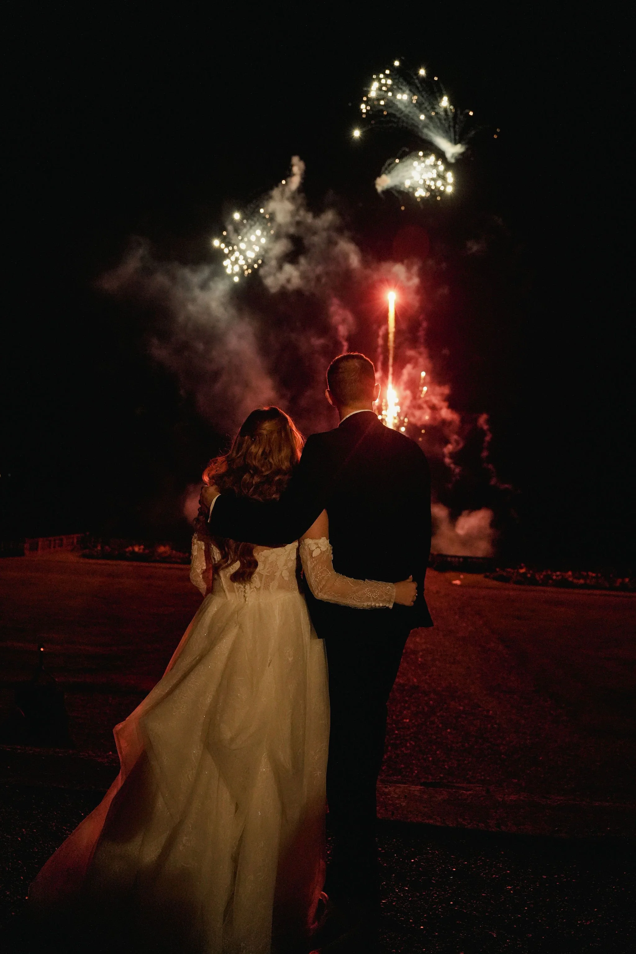 A couple, dressed in formal wedding attire, stands outdoors at night watching fireworks explode in the sky.