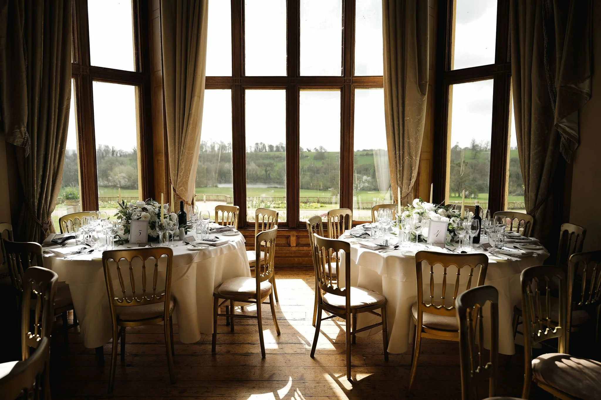 Restaurant dining area with round tables draped in white cloths and beige chairs, set with glassware, silverware, and floral centerpieces. Large window with wooden framing and cream curtains overlooks a green landscape with trees and hills.