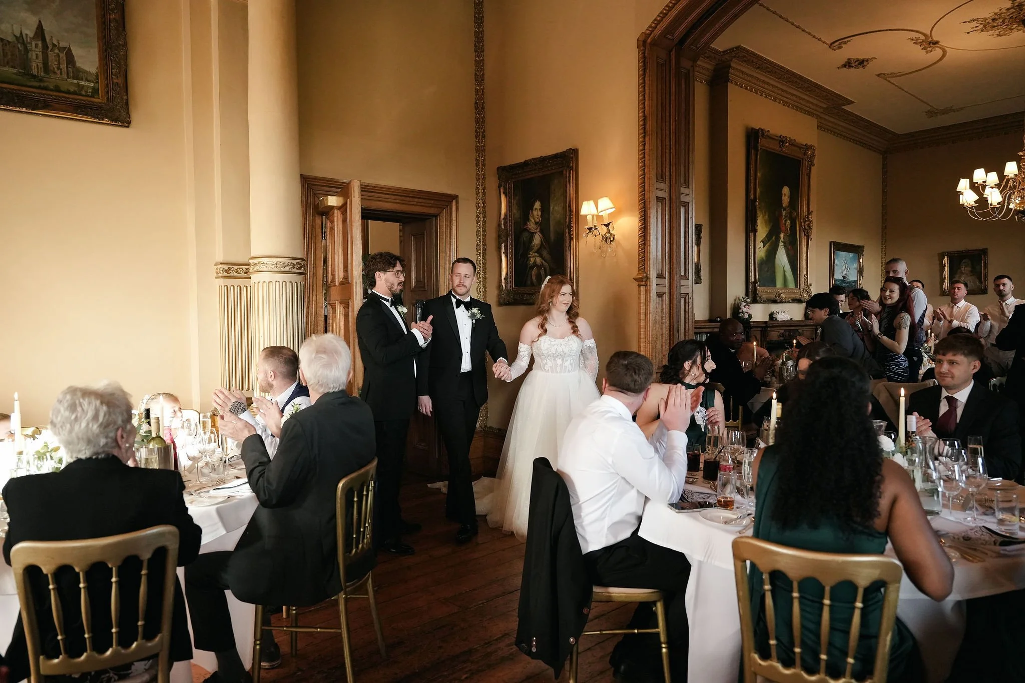 A wedding reception with guests seated at tables, a bride and groom holding hands at the front, and people clapping and watching the couple. The room has ornate woodwork, large framed paintings, and elegant chandeliers.