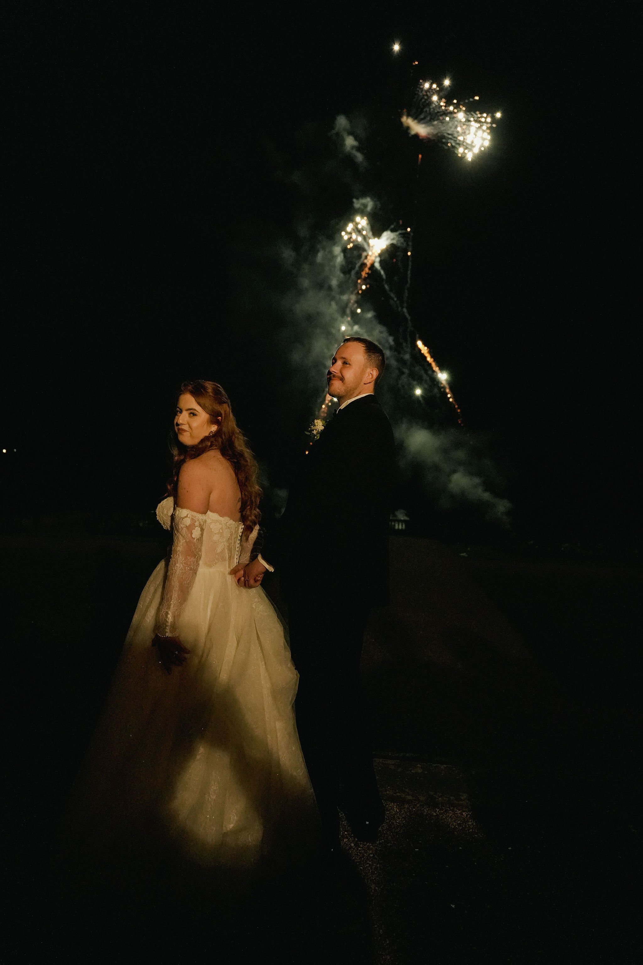 A bride and groom holding hands at night, with fireworks in the sky behind them.