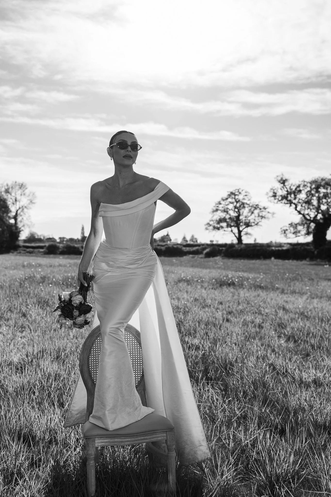 A woman in a wedding dress standing outdoors in a grassy field. She is holding a bouquet of flowers and wearing sunglasses. The sky is partly cloudy with trees in the background.