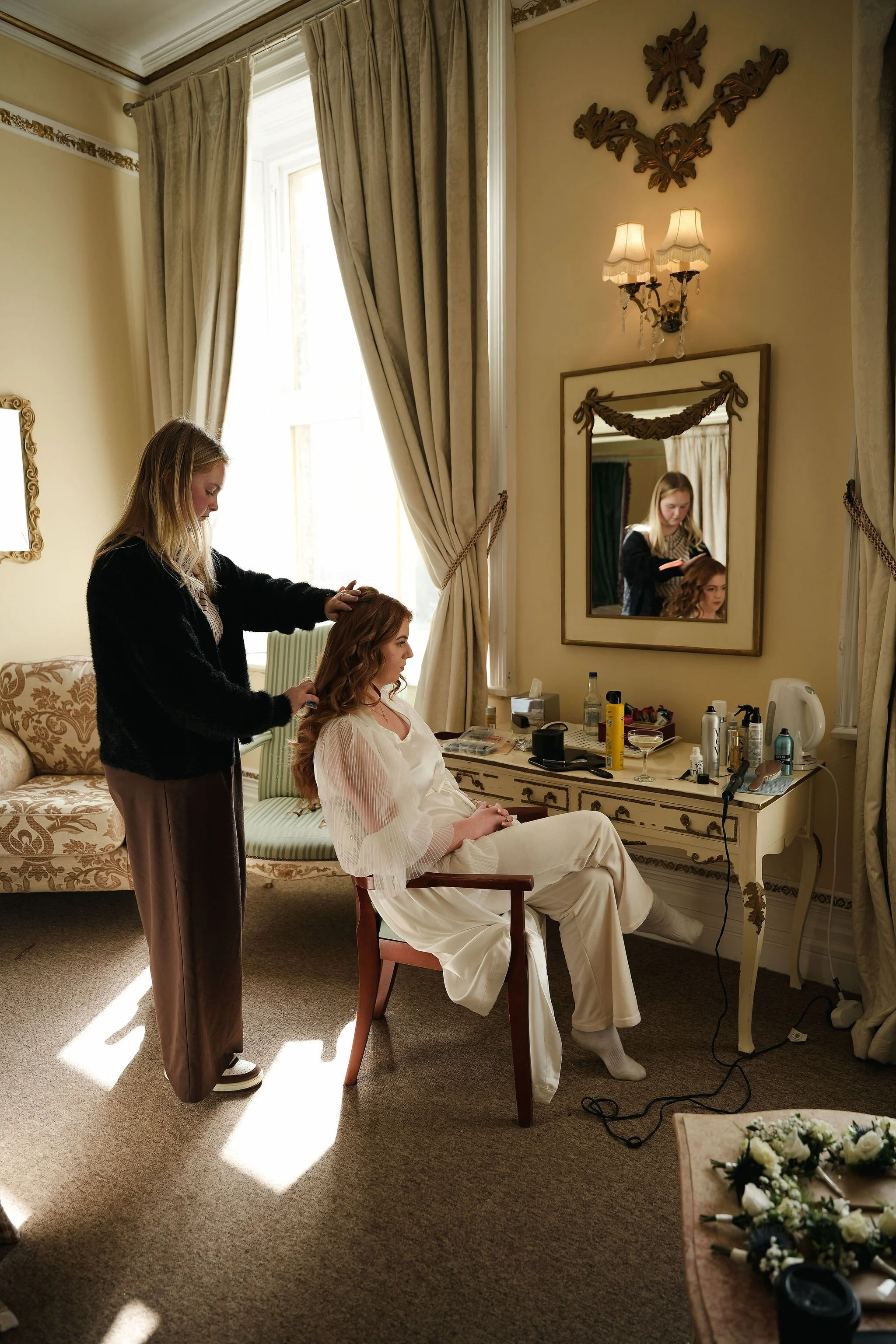 A woman gets her hair styled by a stylist in a luxurious, vintage-style room. The stylist stands behind her, working on her hair, while the woman sits in a wooden chair dressed in a white satin outfit. The room has large windows with elegant curtains