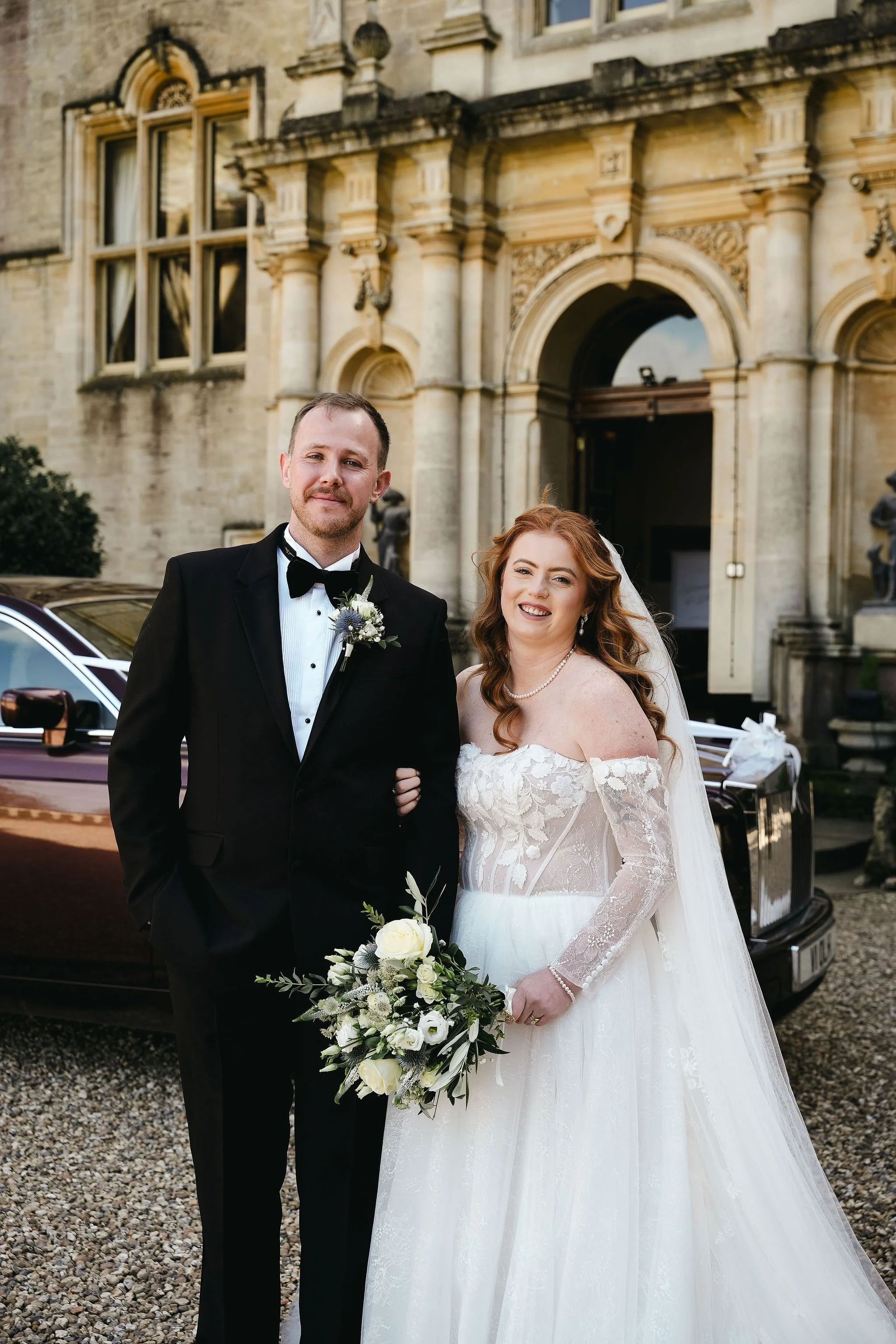 A bride and groom standing outside a historic building with ornate architecture. The groom is dressed in a black tuxedo with a bow tie, and the bride is in a white lace wedding gown holding a bouquet of white and green flowers. They are smiling at th