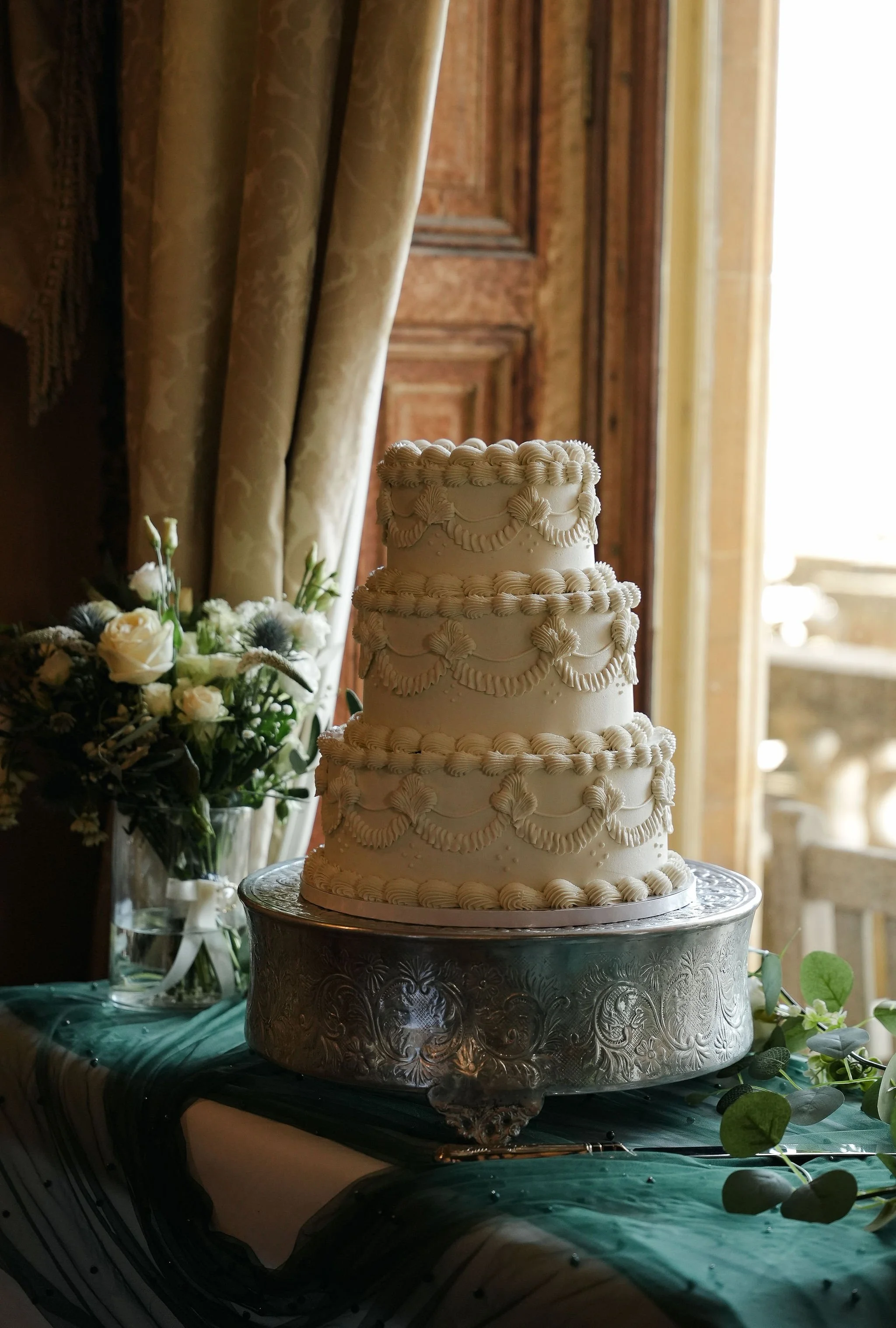 A multi-tiered wedding cake decorated with piped icing patterns on a silver cake stand, placed on a table with greenery and flowers, with curtains and wooden paneling in the background.