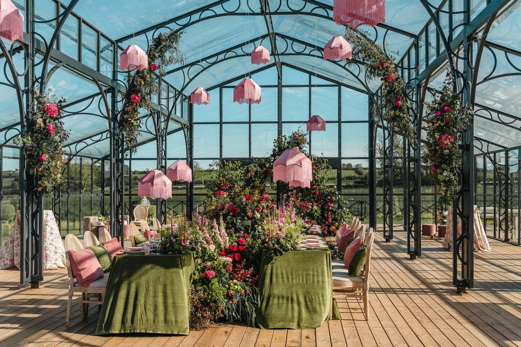 A greenhouse decorated for a wedding with pink parasol lanterns hanging from the ceiling and floral arrangements along the walls. There are tables and chairs set up for guests, with pink and floral-themed table settings and pillows.