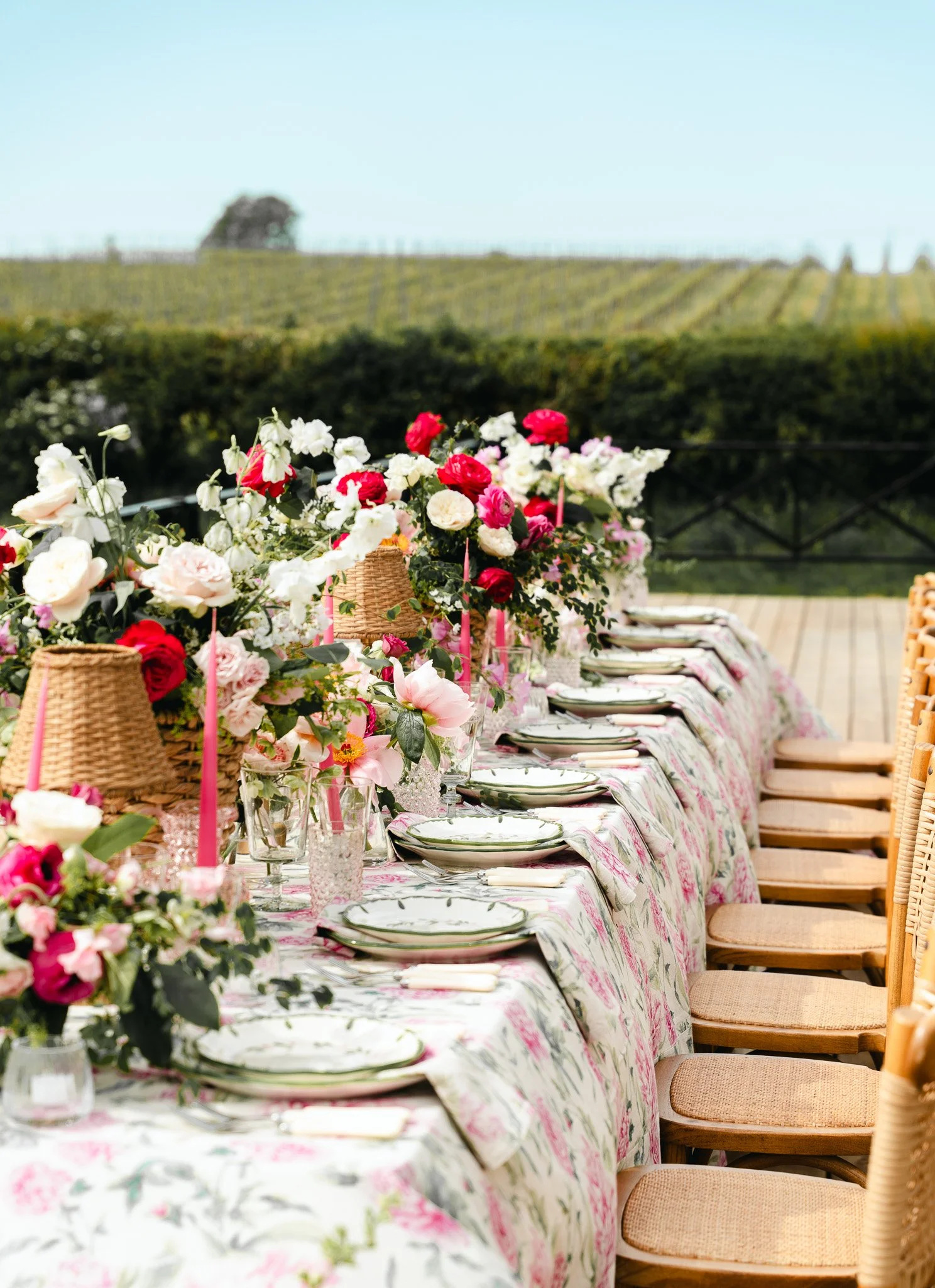 A long table set for a meal outdoors under a clear sky, decorated with colorful flower arrangements, candles, and tableware, with a vineyard and a tree in the background.