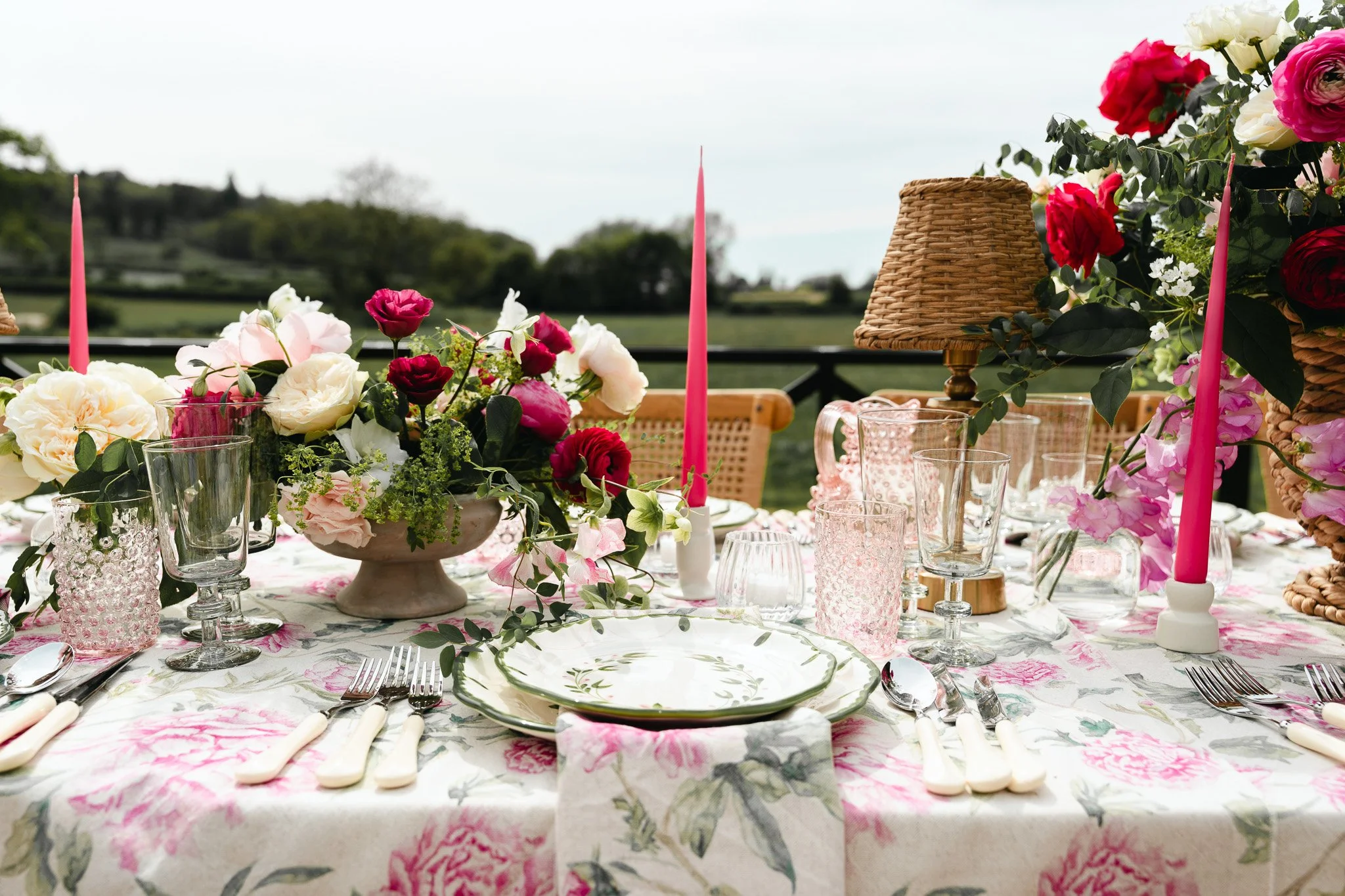 A decorated outdoor dining table with floral arrangements, pink candles, glassware, and a floral tablecloth with a scenic background.