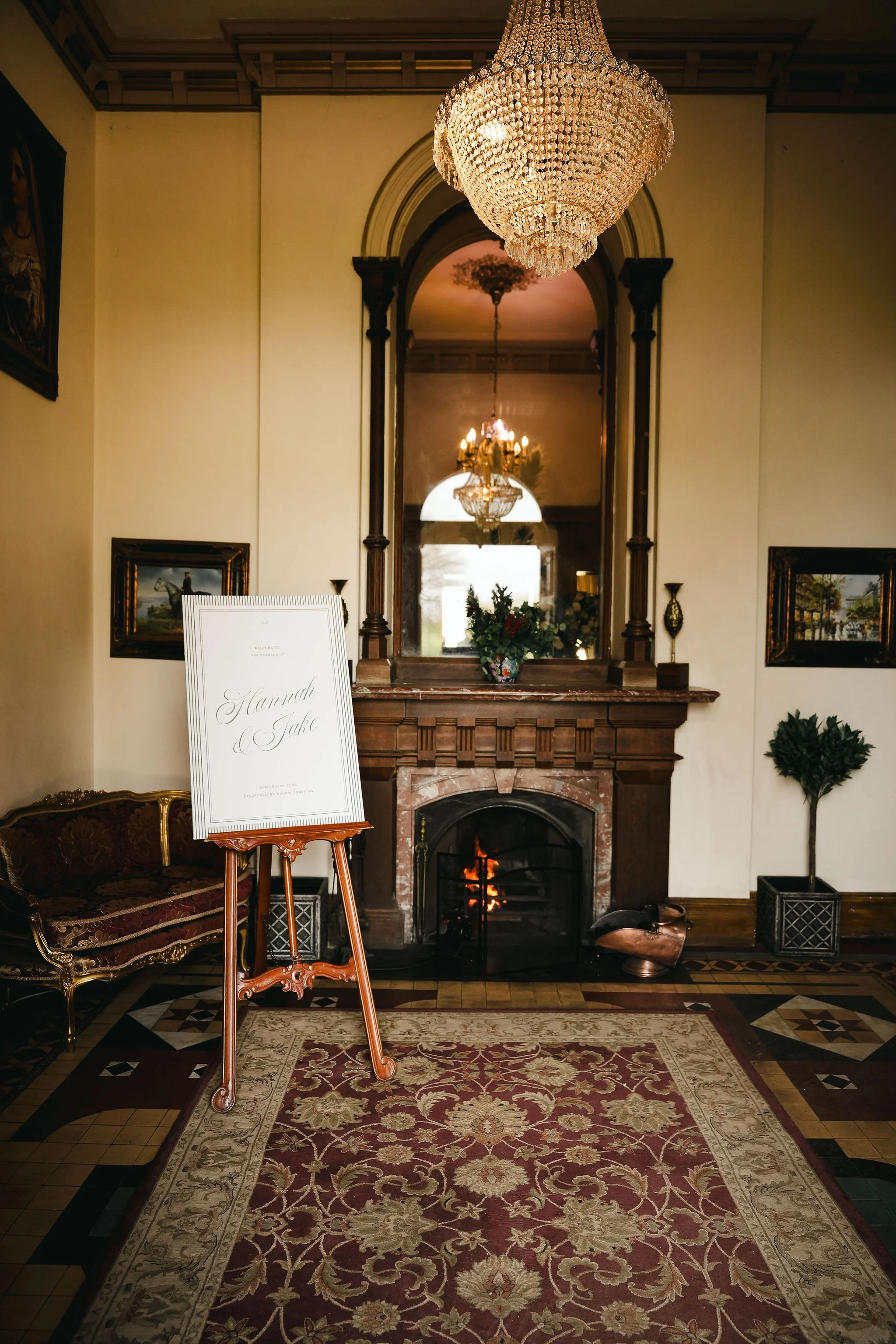 Elegant indoor room with a brick fireplace, ornate chandelier, framed paintings, potted plants, and a rug, featuring a wedding sign for Hannah and Jake.