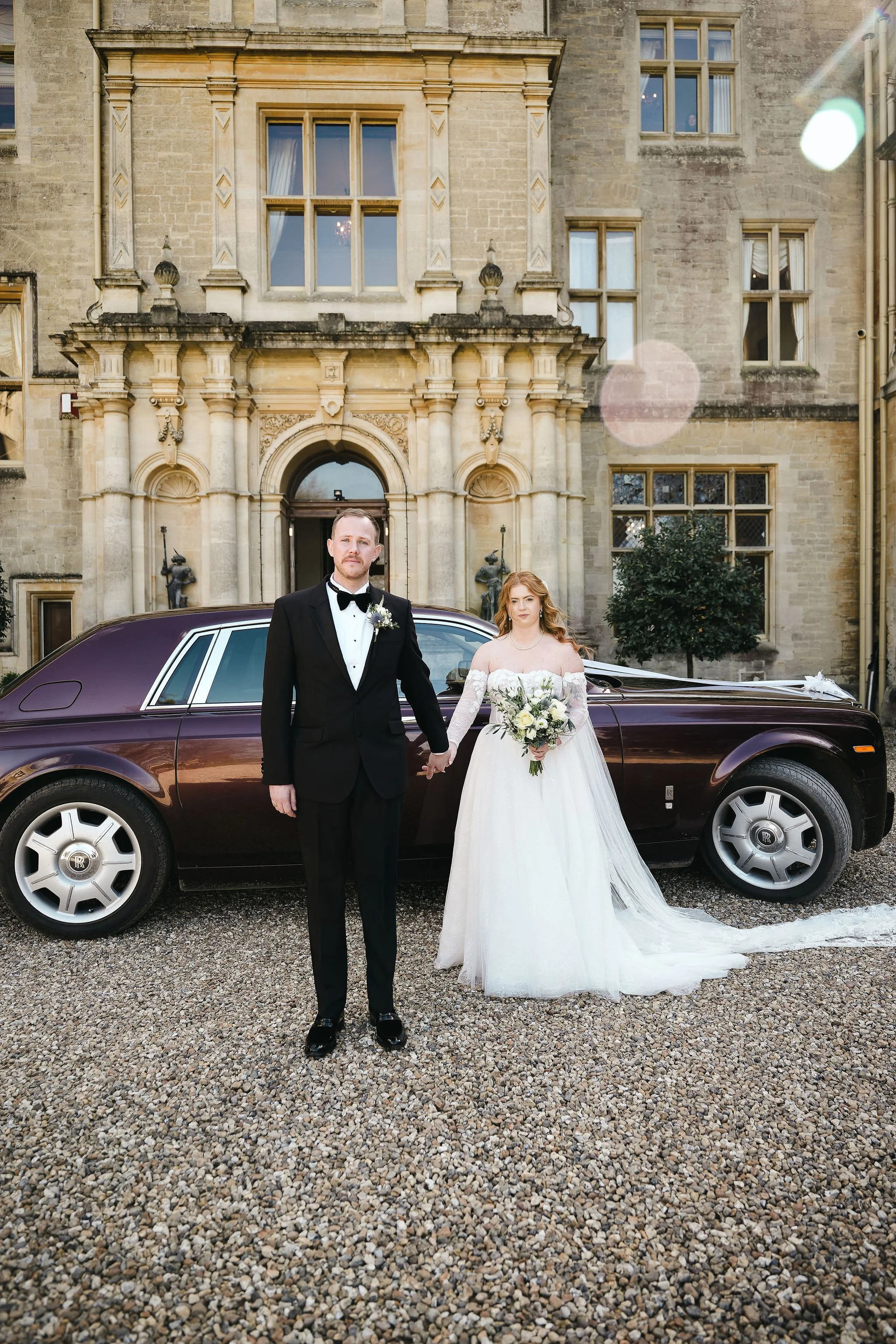 A bride and groom standing in front of a vintage car outside a historic stone building. The groom is wearing a tuxedo with a bow tie, and the bride is in a white wedding gown holding a bouquet of flowers. They are holding hands and looking at the cam