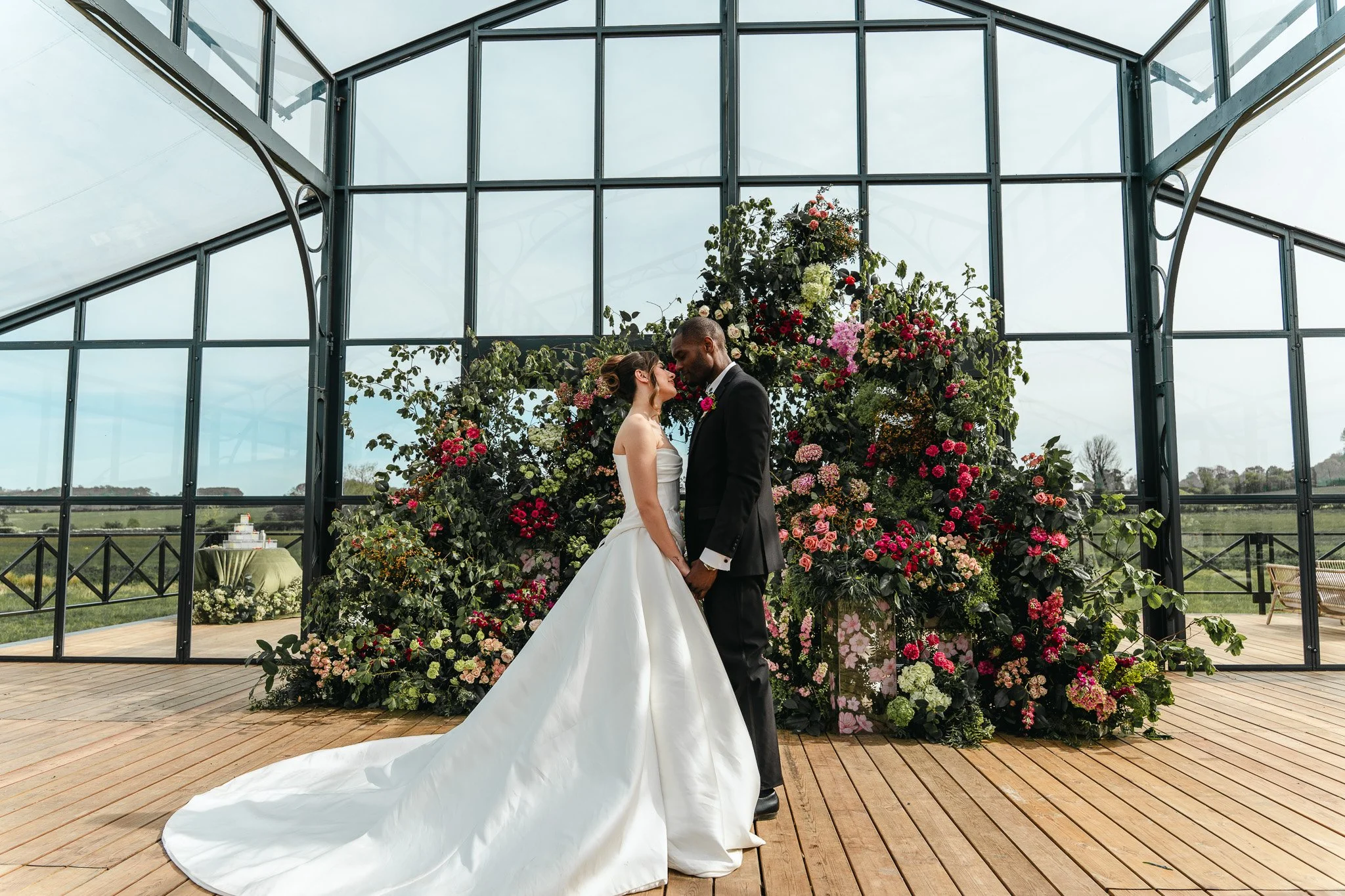 A bride and groom holding hands and looking into each other's eyes during their wedding ceremony, with a large floral arrangement and glass greenhouse structure behind them.