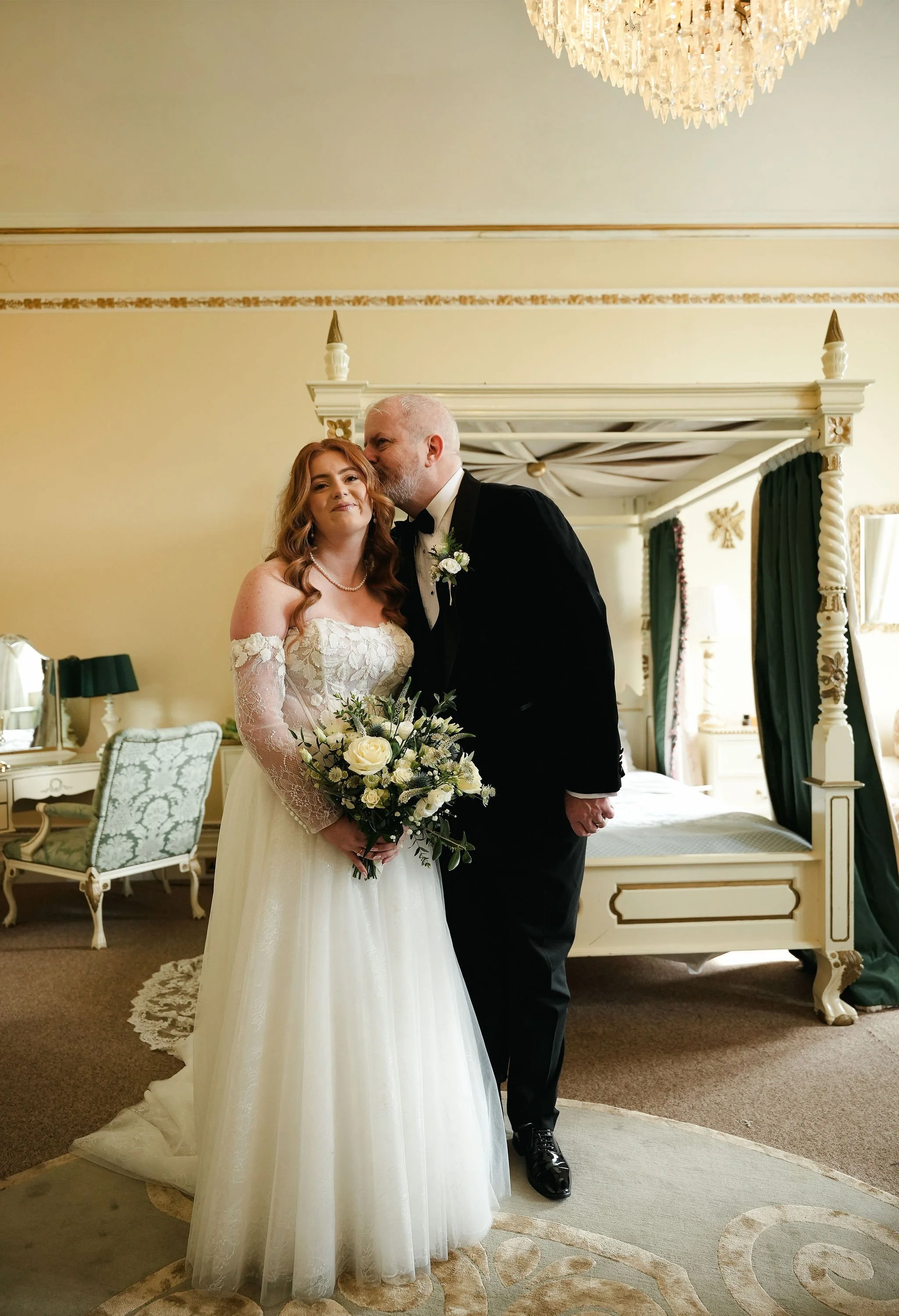 A bride and an older man, likely her father, sharing a kiss on the cheek during a wedding in a decorated room with canopy bed, green curtains, and elegant furniture.
