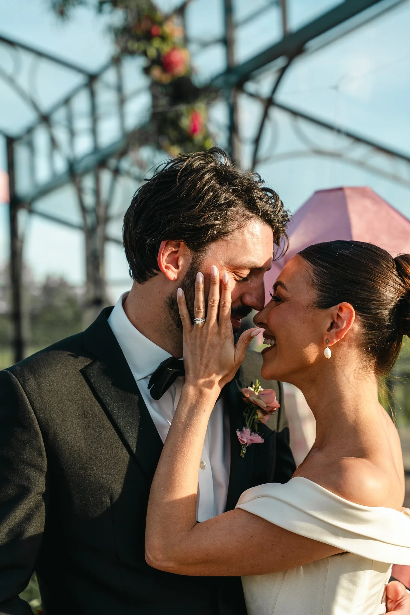 A bride and groom sharing a close, intimate moment during their wedding, with a metal structure and a pink umbrella in the background.