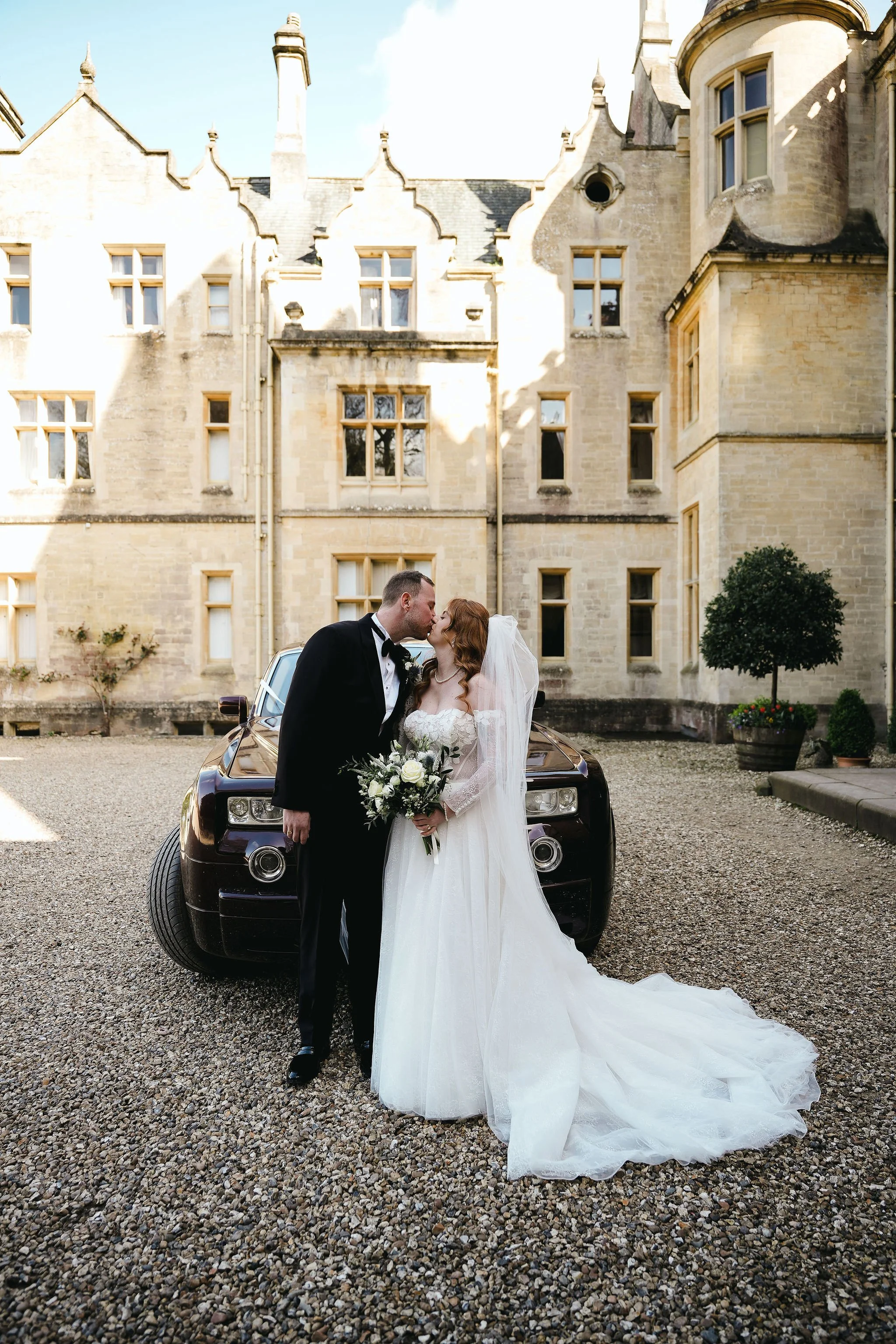 A newlywed couple sharing a kiss in front of a vintage black car, with an old stone building with tall windows and intricate roof details in the background.