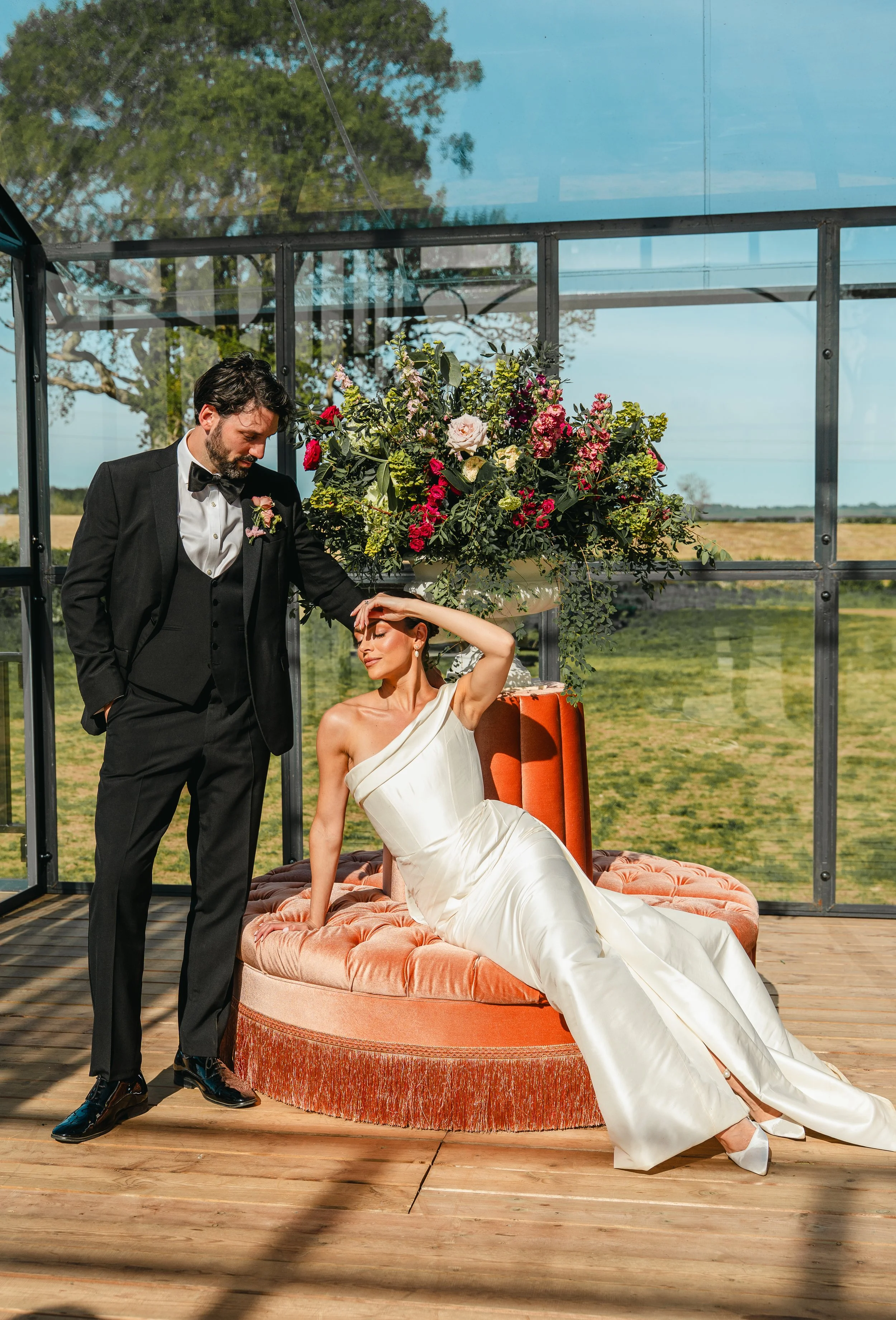 A man in a black tuxedo standing beside a woman in a white gown who is sitting on an orange velvet upholstered bench, inside a glass enclosure with a large floral arrangement above her head, on a wooden platform outdoors.
