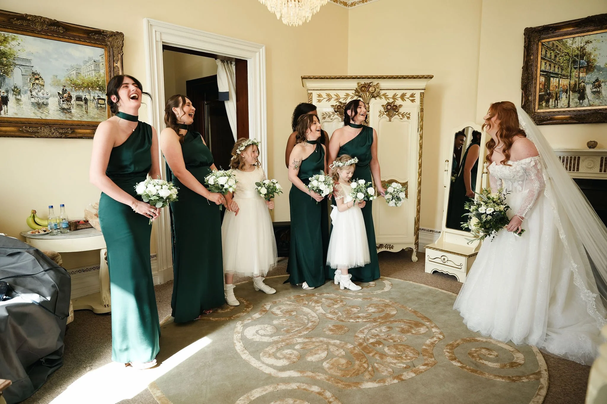 Bride with long red hair in wedding dress smiling at bridesmaids and flower girls, who are laughing and holding bouquets, in a decorated room.