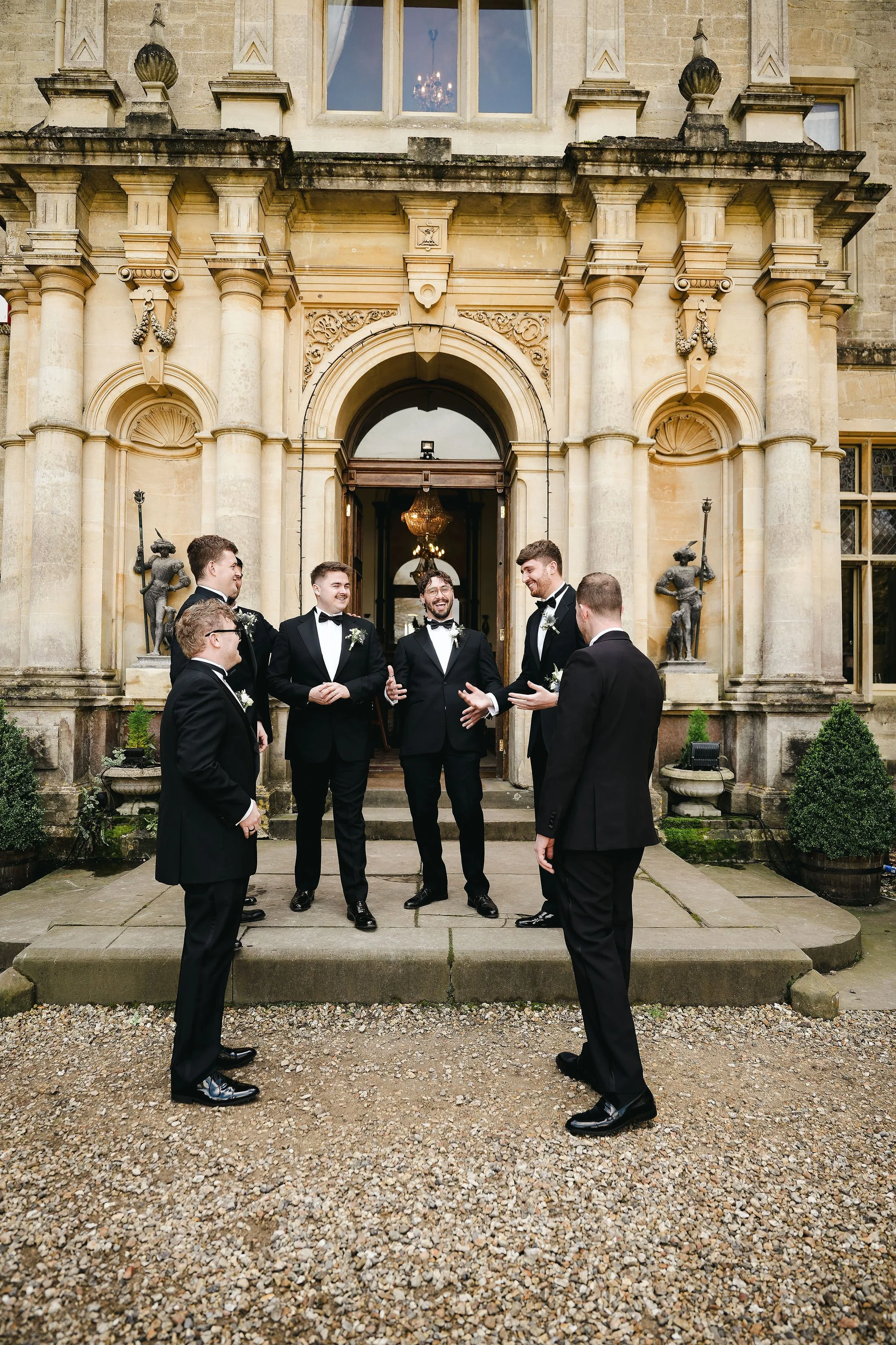 Groom and Groomsman dressed in black tuxedos standing outside a grand building with stone architecture, engaging in conversation, with a chandelier visible through the open door behind them.