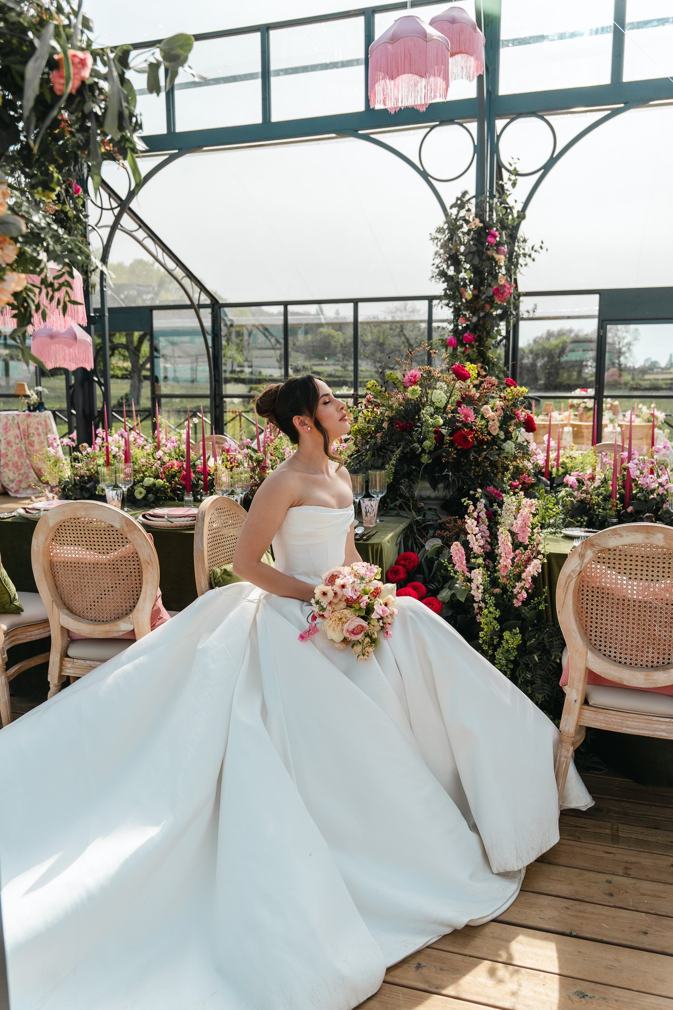 A bride sitting on a bench surrounded by pink and white flowers in a conservatory or greenhouse setting.
