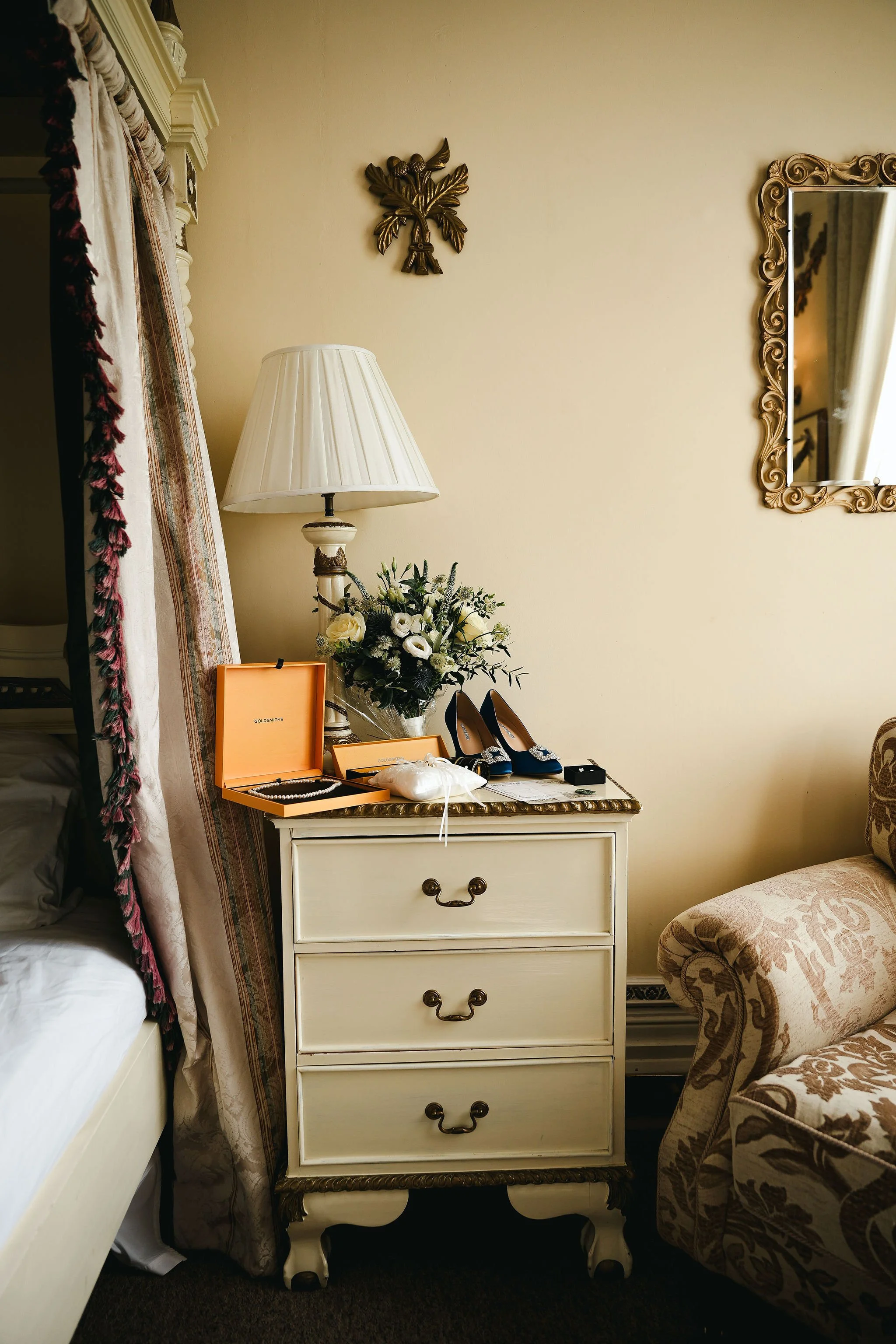 A cream-colored wooden nightstand with three drawers, ornate handles, and curved legs, situated between a bed with white bedding and a floral-patterned armchair, topped with a table lamp, a bouquet of flowers, and jewelry boxes, against a beige wall 