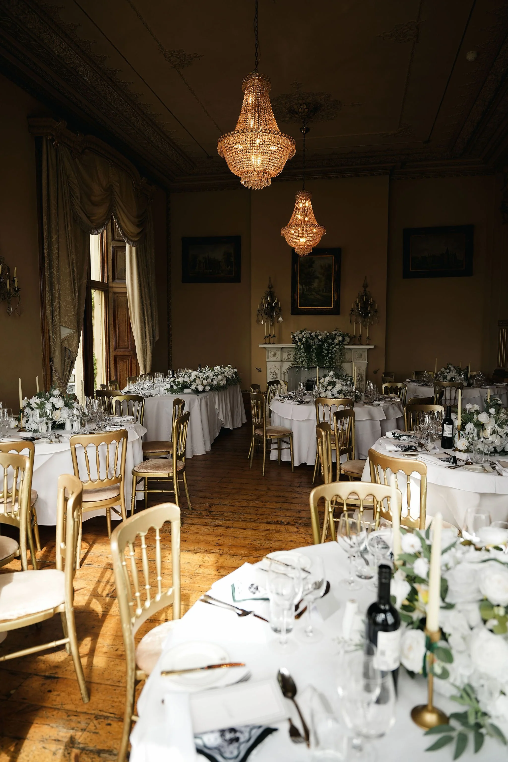 Elegant dining room with round tables covered in white tablecloths, decorated with white flowers, candles, and tableware, gold chairs, ornate ceiling, large chandeliers, framed paintings, fireplace, and large windows with drapes.