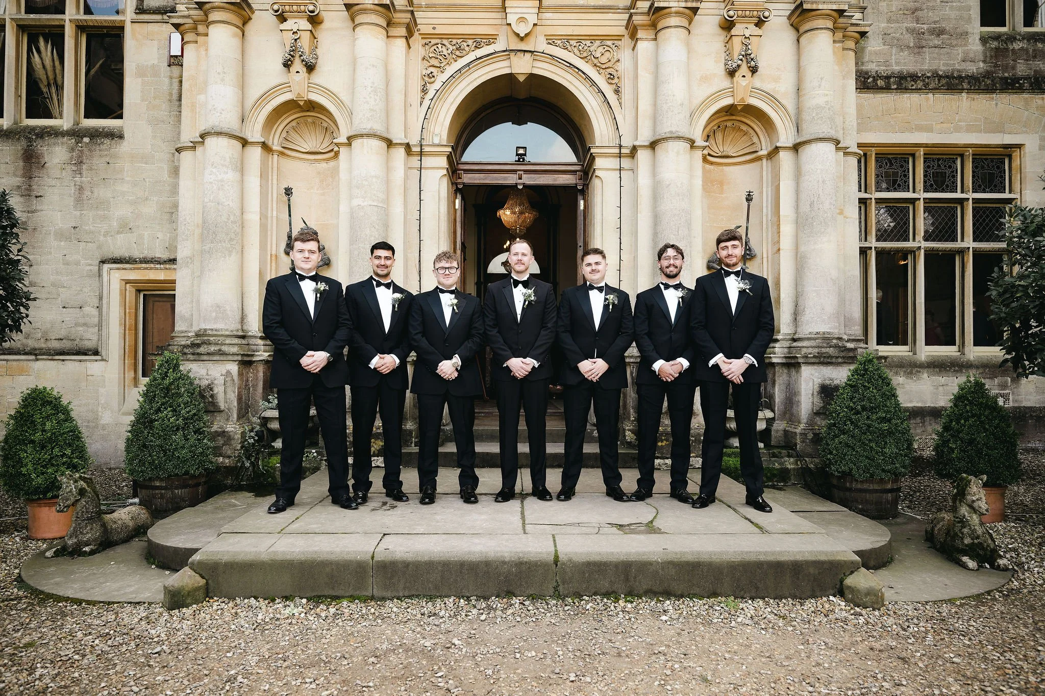 Seven men dressed in tuxedos standing in front of a large historic building with ornate columns, a chandelier visible through the open door, and potted plants on either side of the entrance.