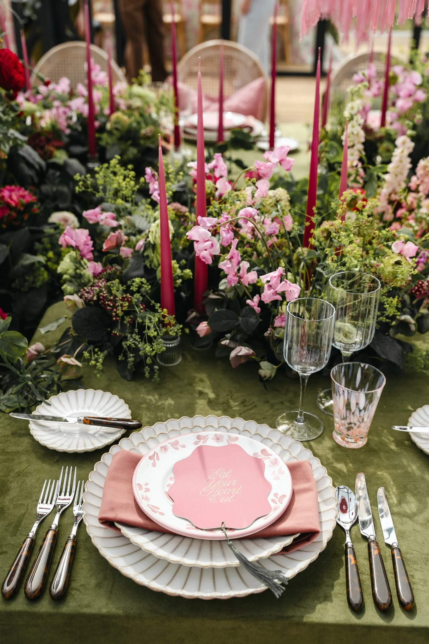 Elegant dining table set with pink and white floral arrangements, red candles, and place settings with pink and white plates, glasses, silverware, and a pink napkin, for a romantic or special occasion.