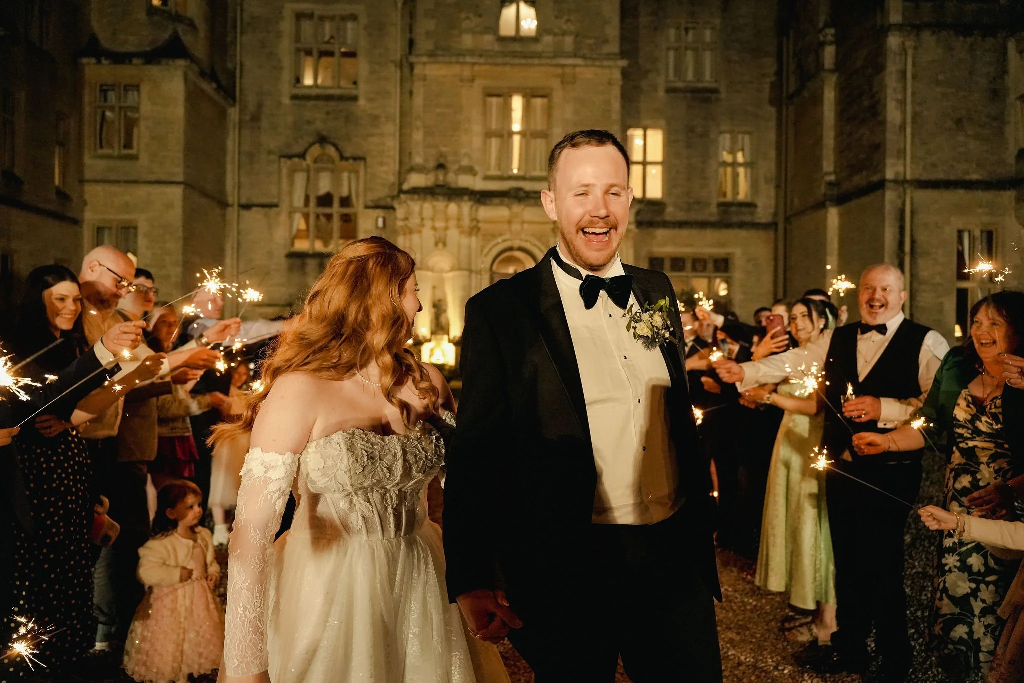 A wedding celebration taking place outdoors at night, with a bride and groom holding hands and walking through a crowd of guests holding sparklers, in front of a large historic building with lit windows.