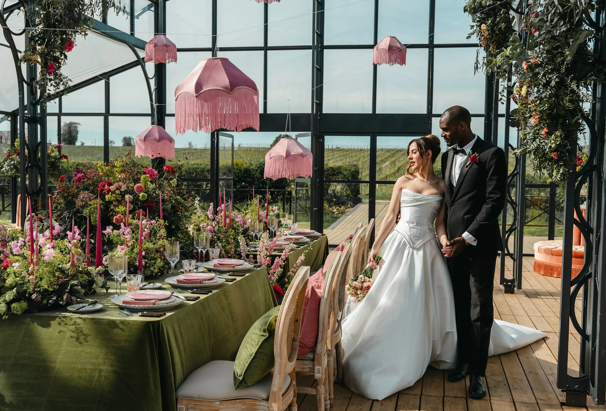 A bride and groom dressed in wedding attire holding hands inside a glass greenhouse decorated with pink lampshades and floral arrangements, overlooking a vineyard landscape.