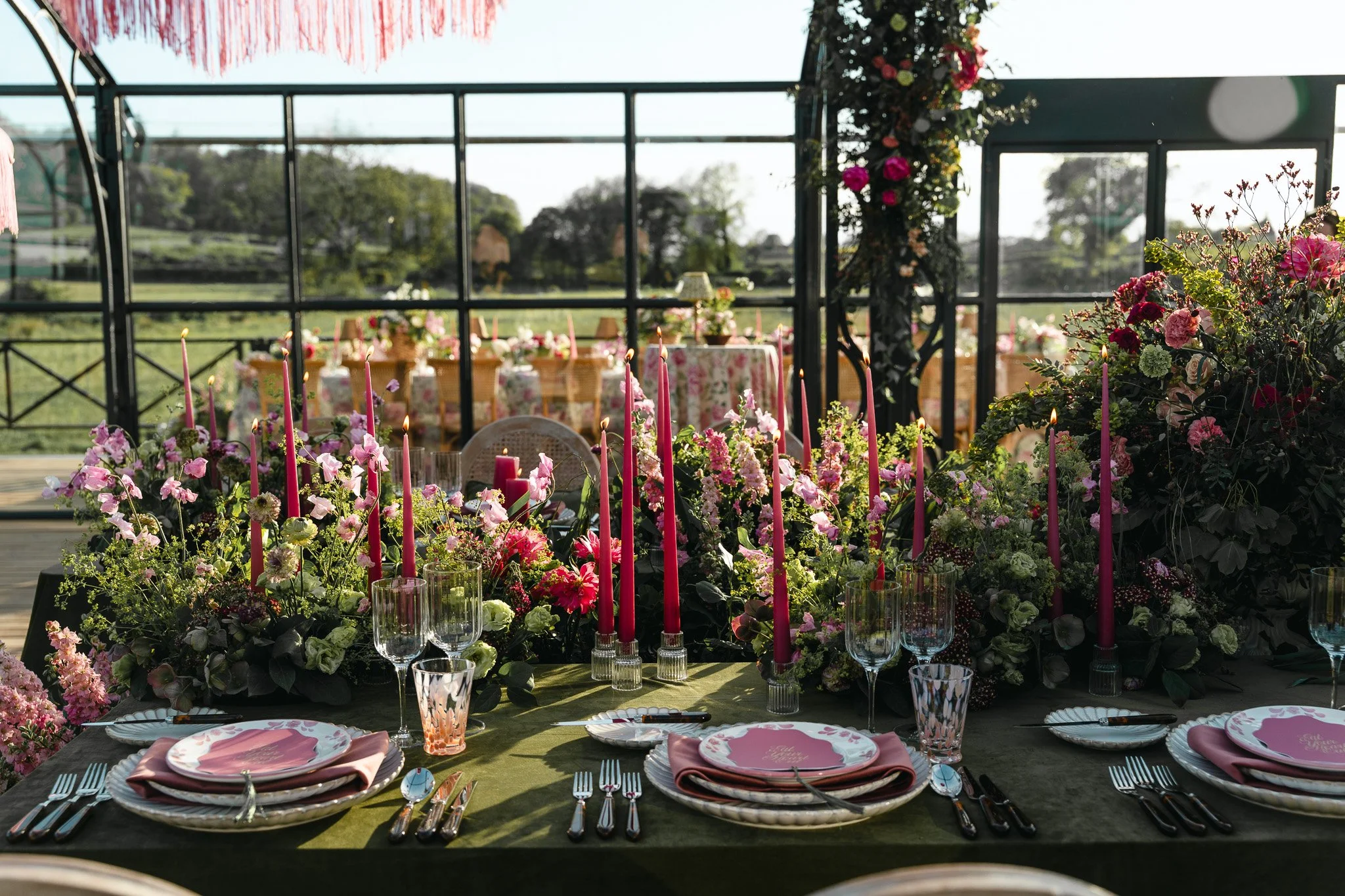 A banquet table decorated with pink candles, flowers, and tableware, set up indoors with large windows showing greenery outside.