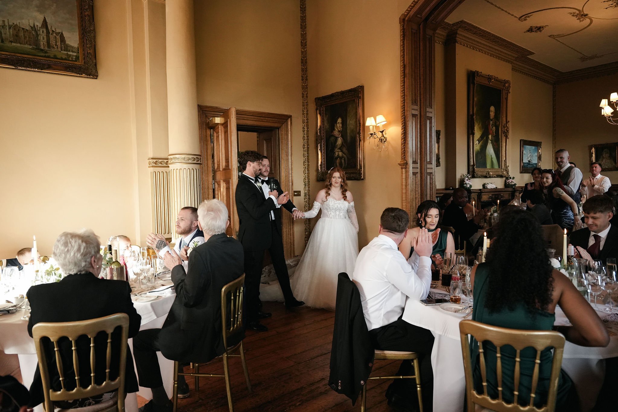 A wedding reception in a grand room with vintage portraits on the wall. The bride and groom are standing together, holding hands. Guests are seated at tables, clapping and watching the couple.
