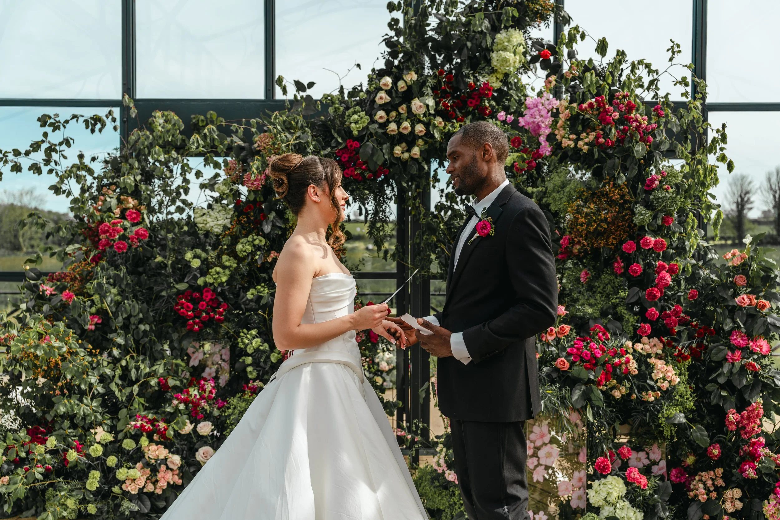 A bride and groom exchanging vows during a wedding ceremony surrounded by a lush floral backdrop of pink, red, and white flowers in a greenhouse or conservatory.