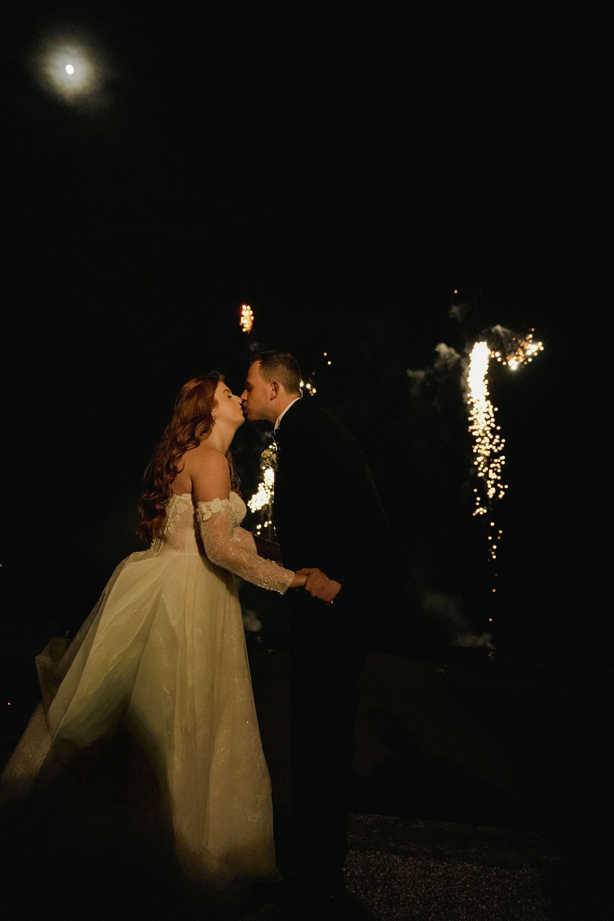 A bride and groom kiss at night with fireworks in the sky behind them.