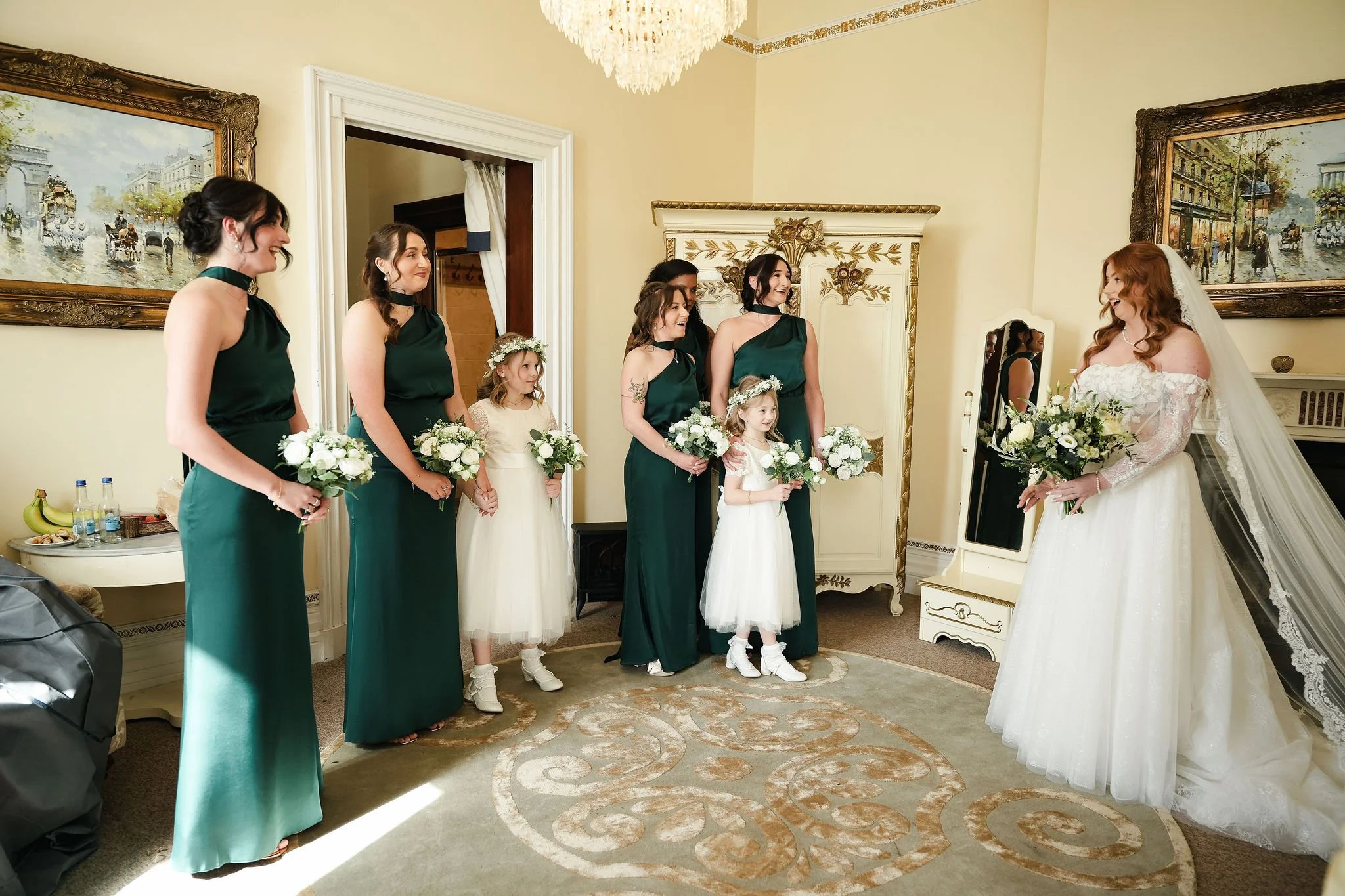 A bride in a white lace wedding dress and veil talking to five bridesmaids and two flower girls, all holding white bouquets, in a decorated room with artwork and a chandelier.