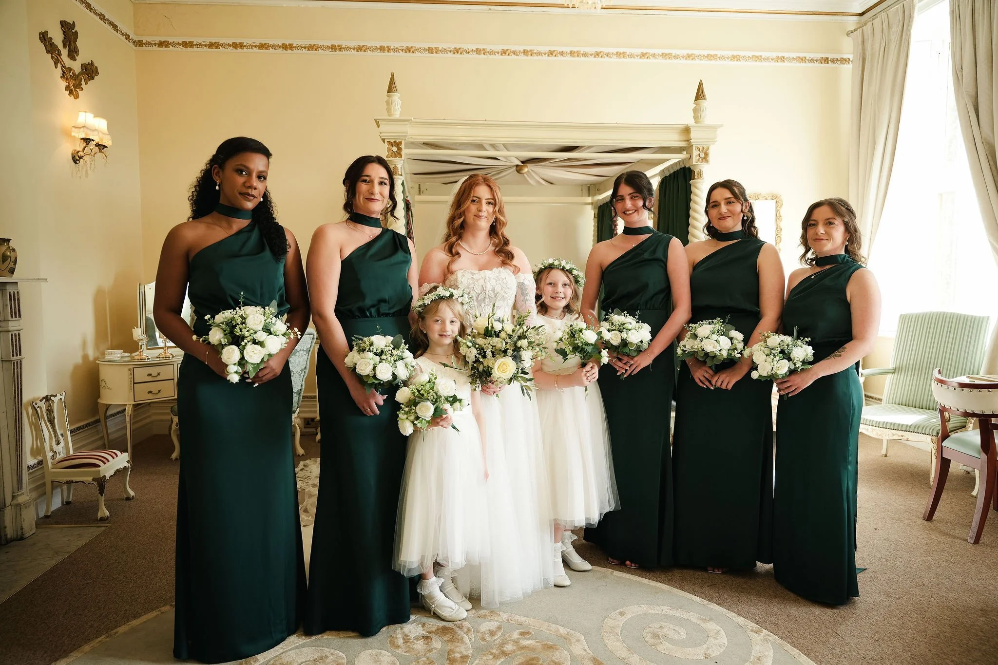 A group of women, likely bridesmaids, standing indoors in a wedding setting, holding bouquets of white flowers and dressed in dark green bridesmaid dresses, with a bride in a white wedding dress and veil in the middle, smiling for a photo.