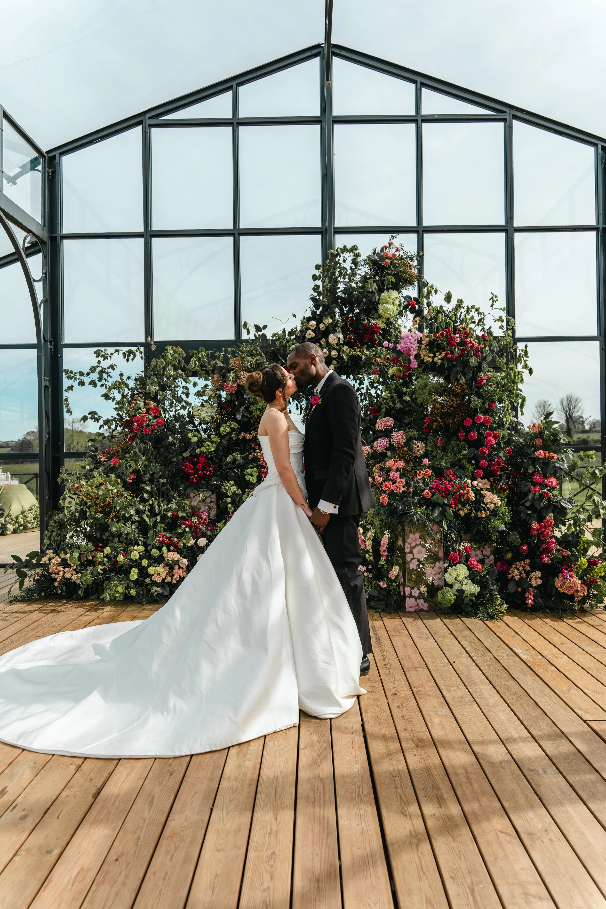 A bride and groom are kissing in front of a floral backdrop at a wedding venue, with a glass greenhouse structure behind them and a wooden floor beneath.