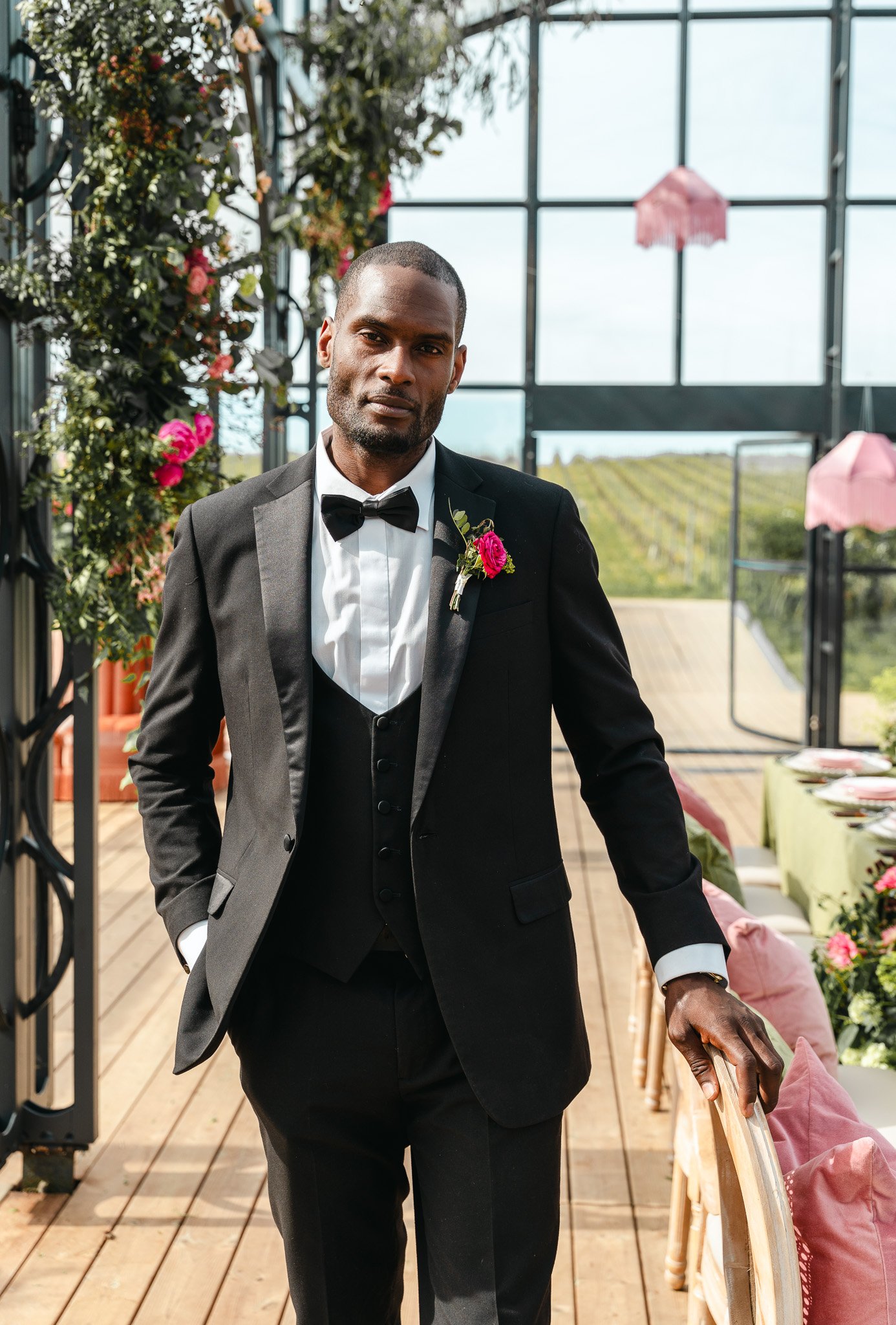 A man in a black tuxedo with a white shirt and black bow tie, wearing a pink flower boutonniere, standing in a sunlit greenhouse with plants and pink pillows on a wooden bench.